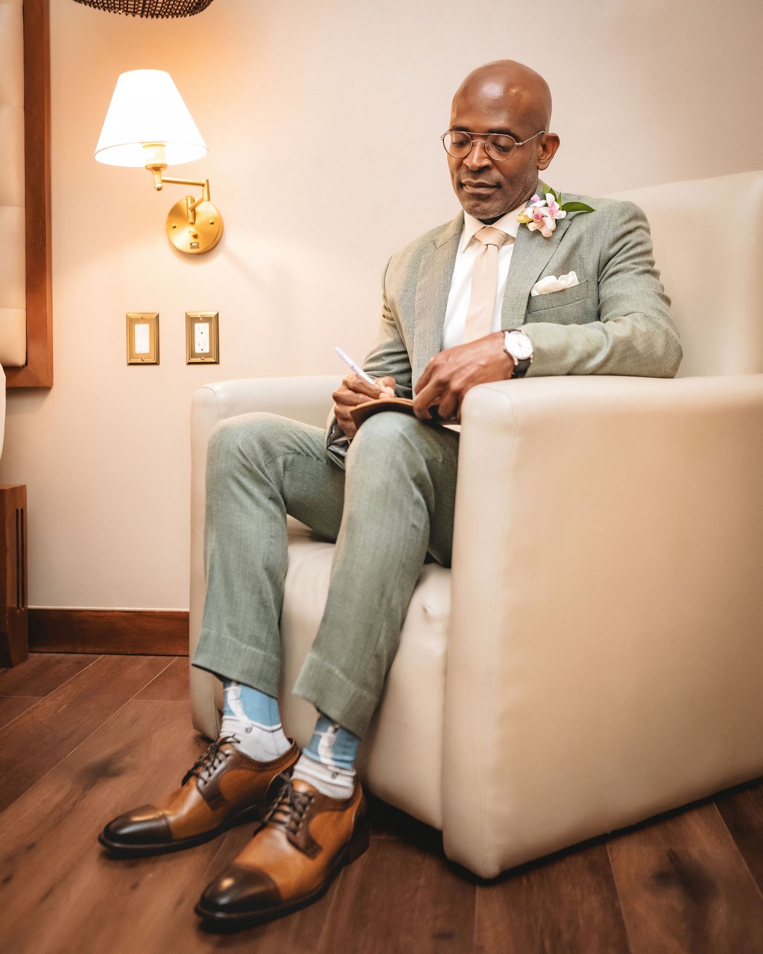 Elegant man in sage suit with floral boutonniere sits in white chair, wearing colorful socks and brown oxford shoes.