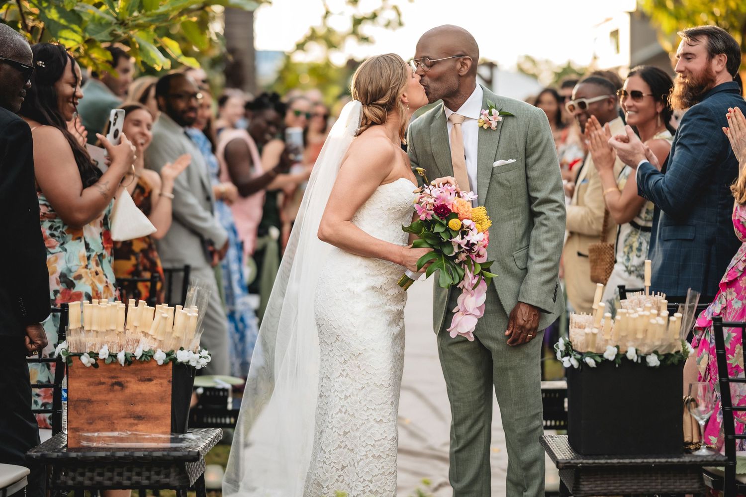 Bride and groom share a kiss at outdoor wedding ceremony surrounded by applauding guests, bride holding colorful bouquet.