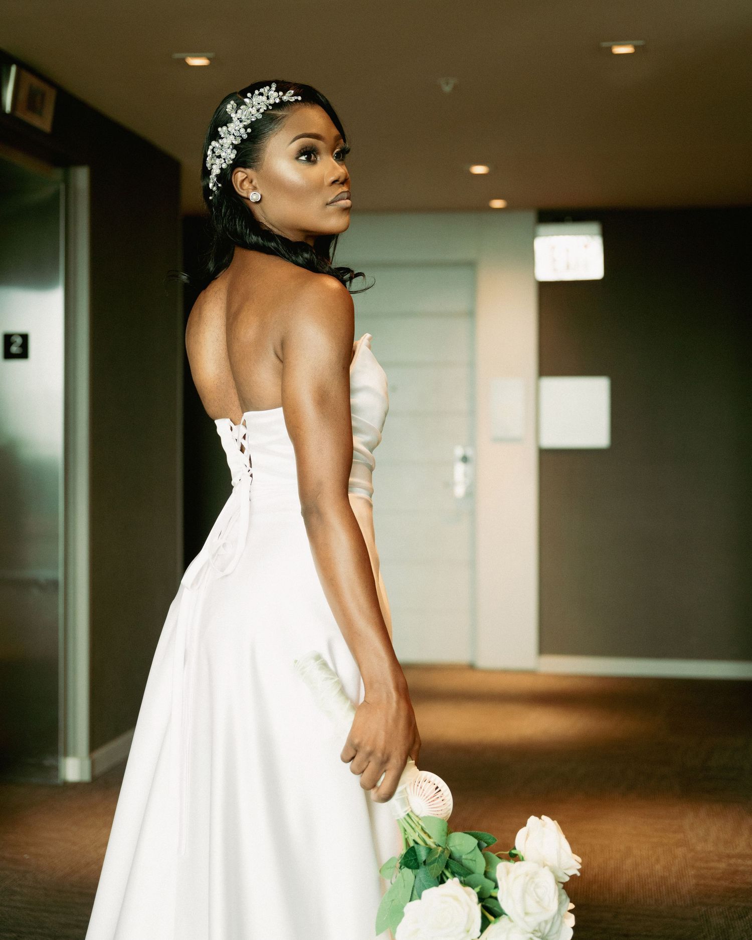 Bride in strapless white wedding gown with crystal headpiece holding pink roses in hotel corridor.