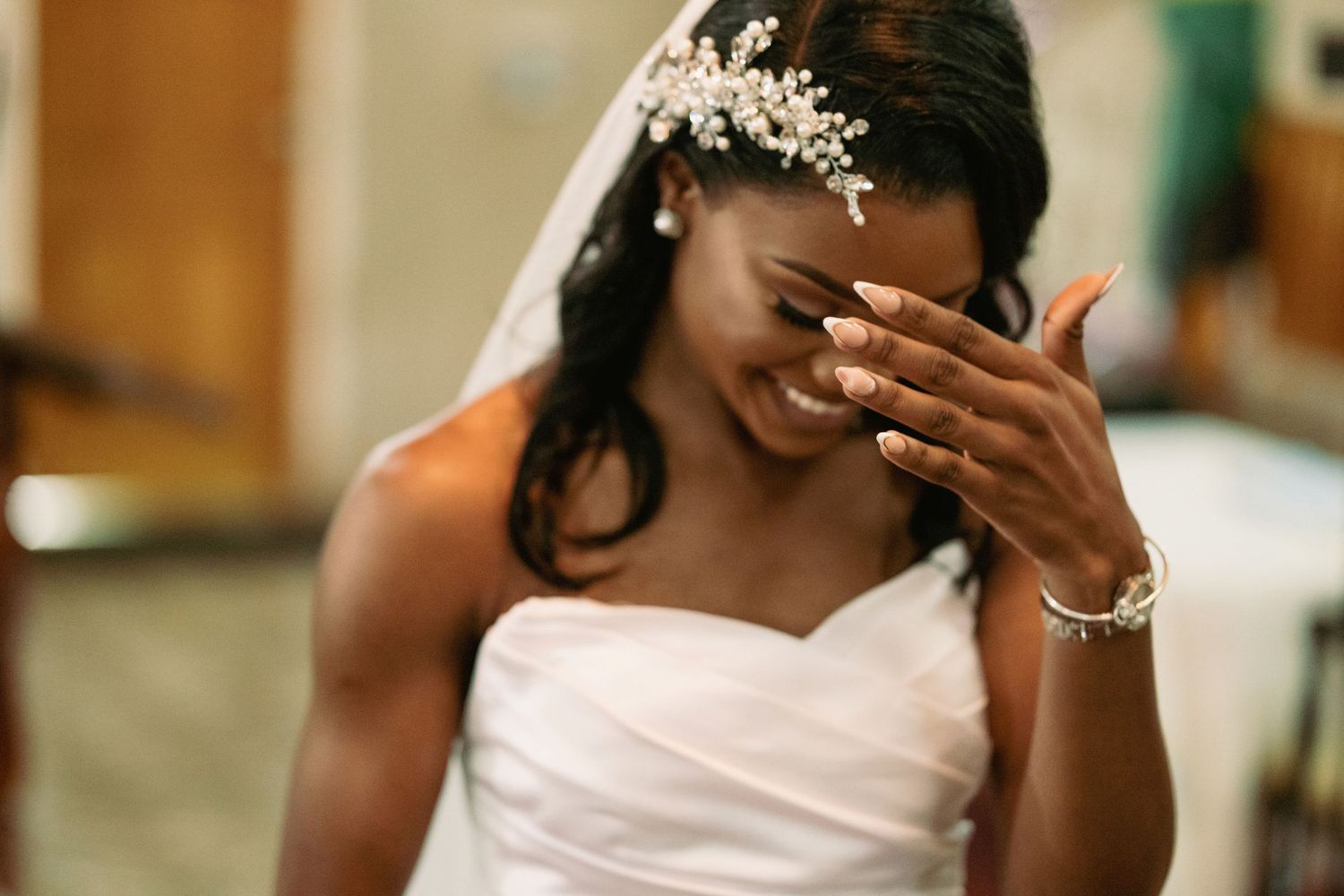 Emotional Black bride in white strapless gown and pearl headpiece covers face while smiling during ceremony.