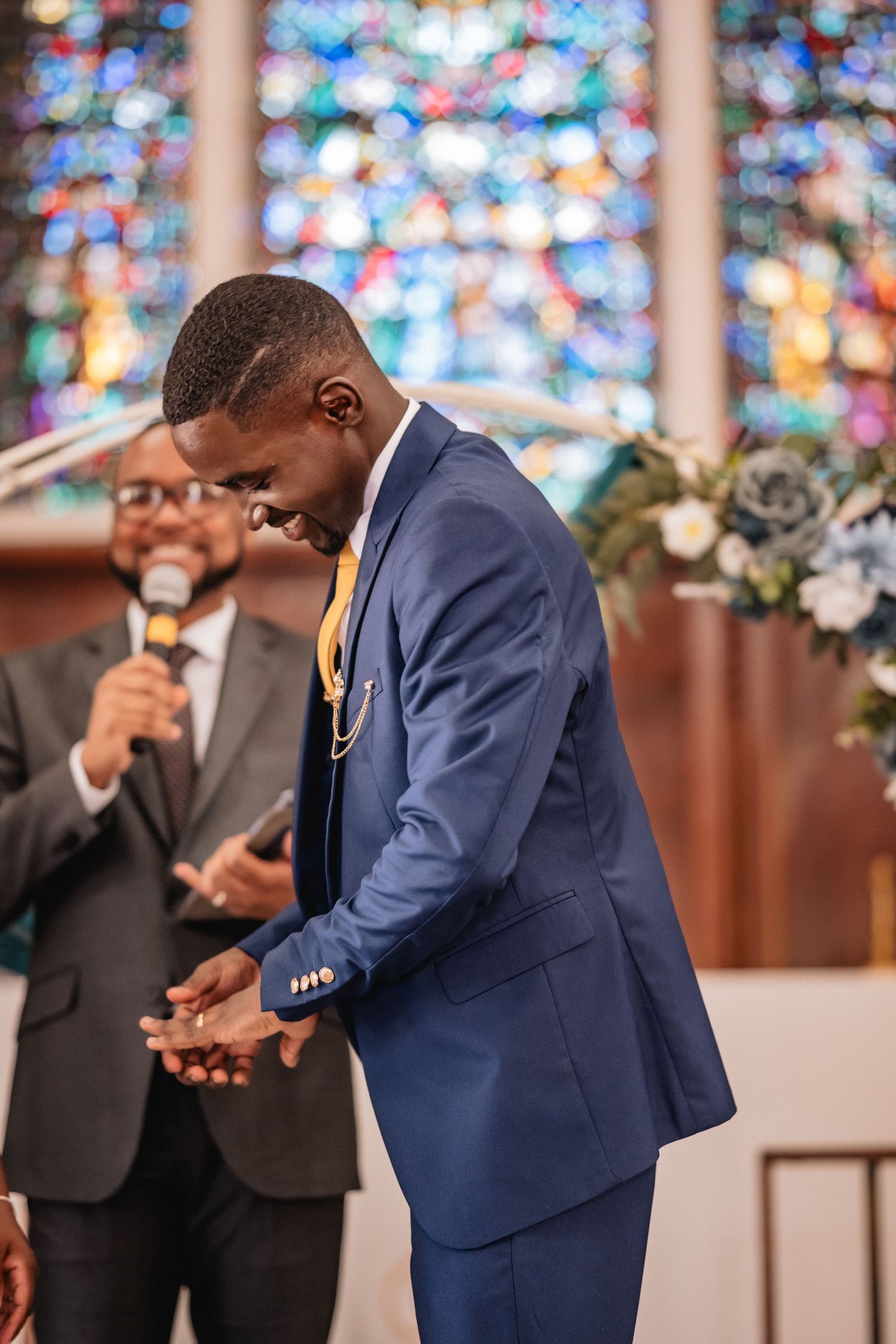 Groom in blue suit laughing joyfully at church altar with colorful stained glass window backdrop during wedding ceremony.