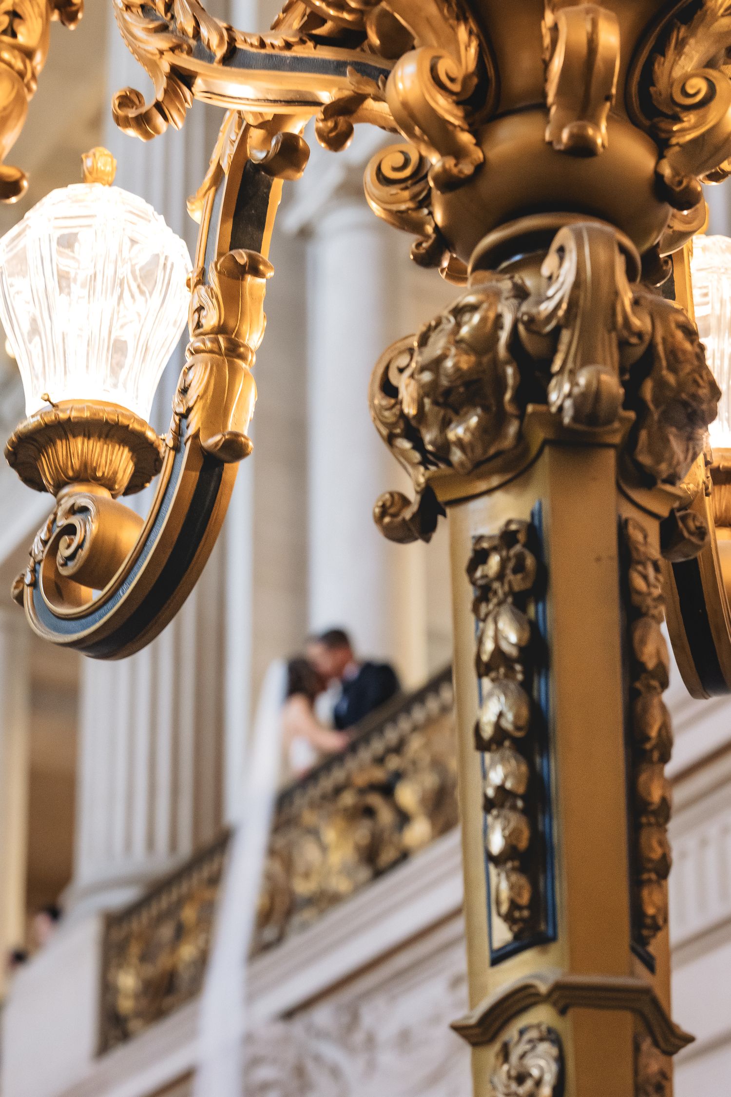 Ornate golden chandelier in elegant ballroom with blurred couple embracing on balcony in background.