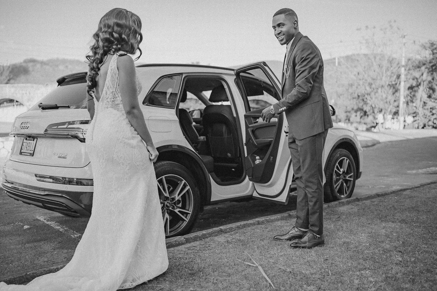 Groom in gray suit opening car door for bride in white wedding dress in black and white photo.