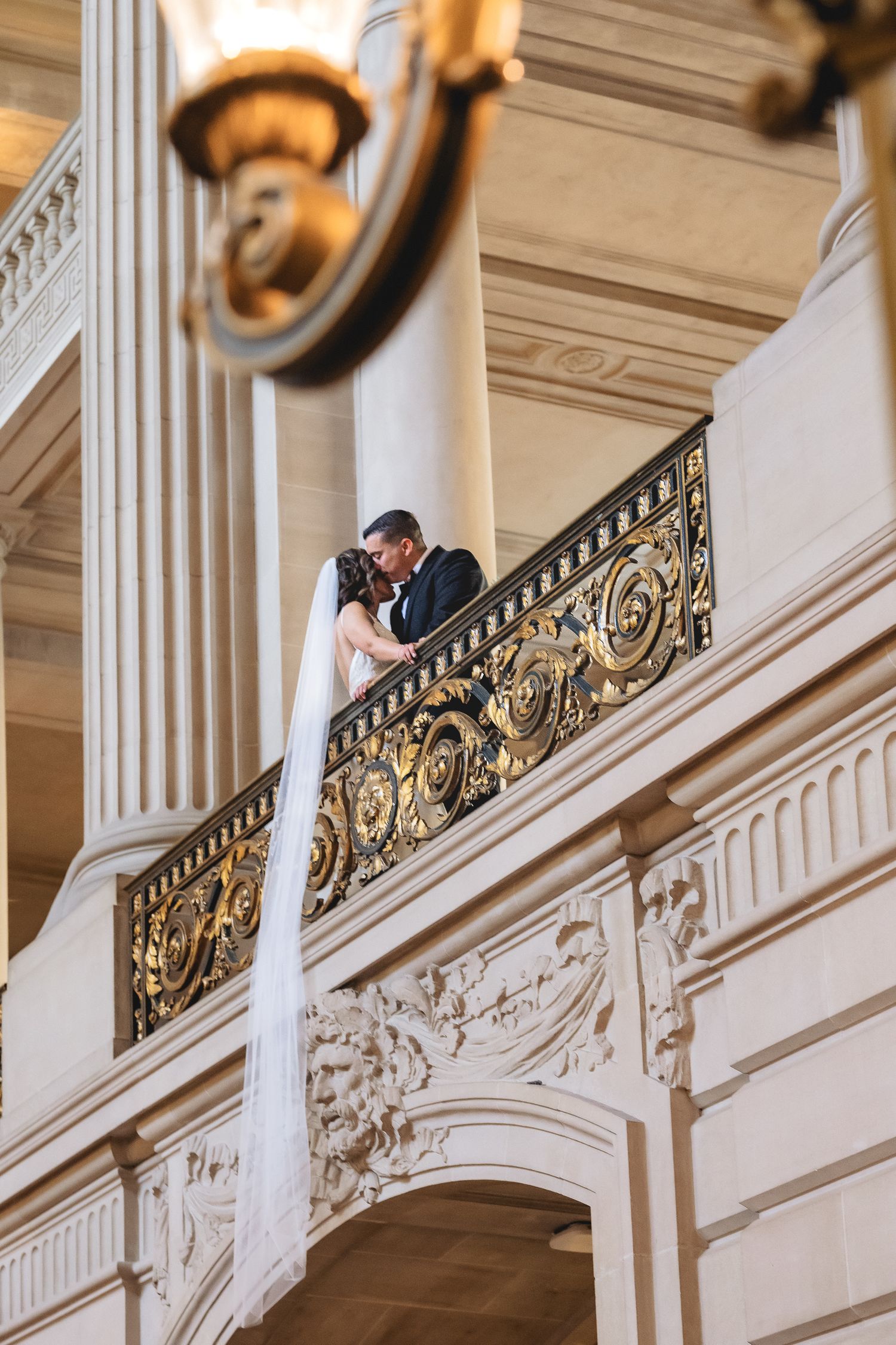 Bride and groom share a romantic kiss on an ornate golden balcony railing inside a grand classical building.