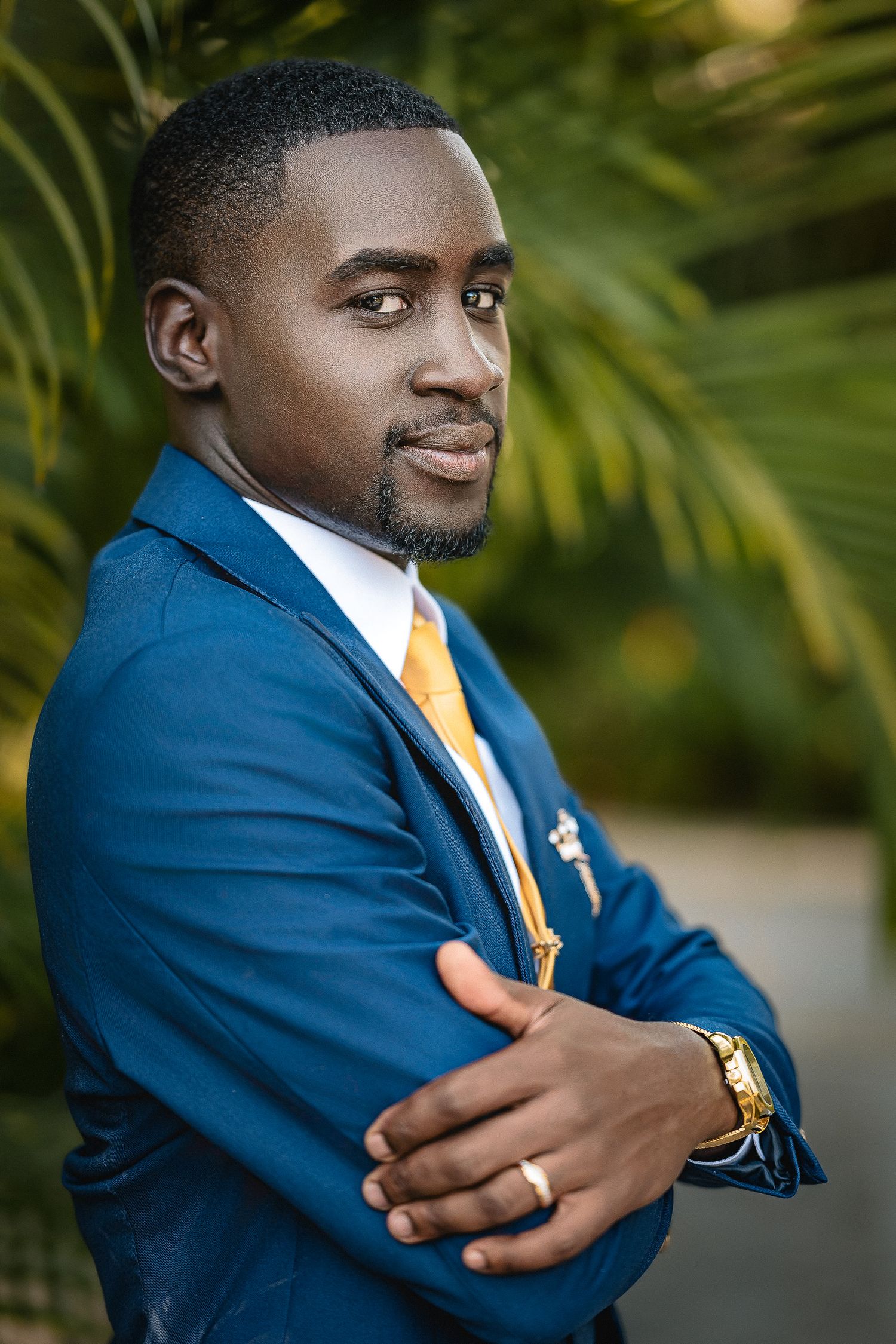 Confident groom in blue suit and gold tie poses with arms crossed among tropical greenery.