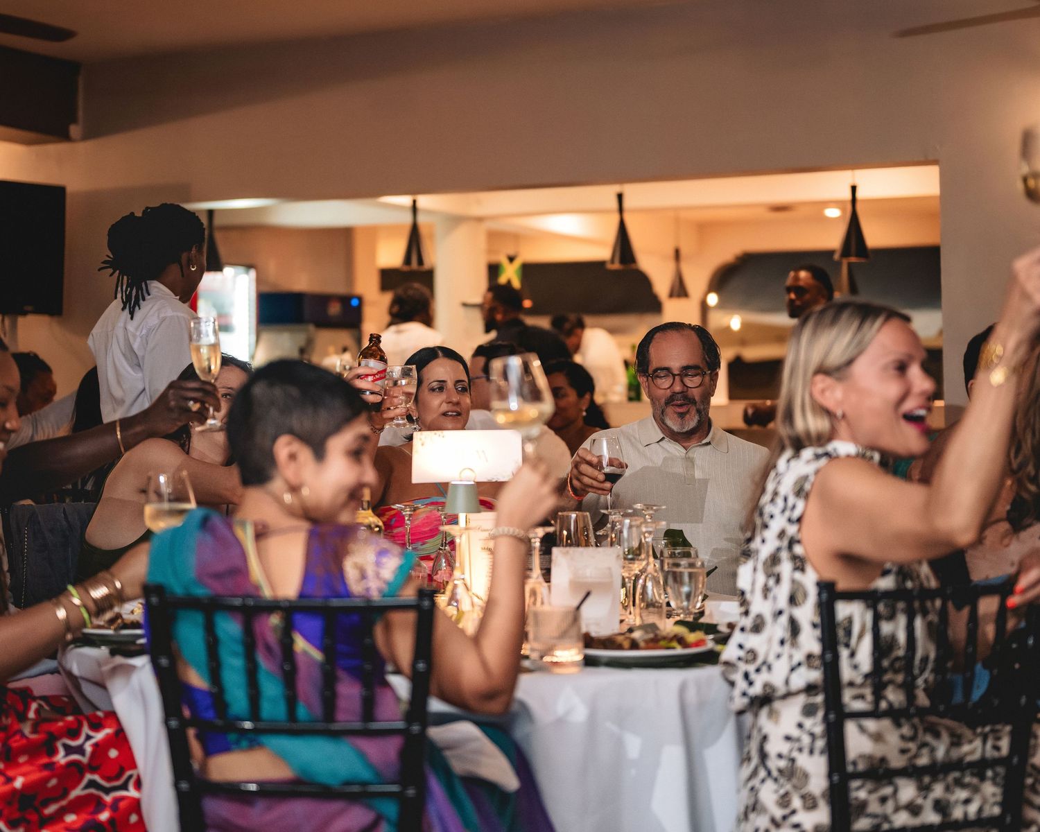 Guests celebrate at a lively indoor dinner party, raising glasses and phones in a warmly lit restaurant.
