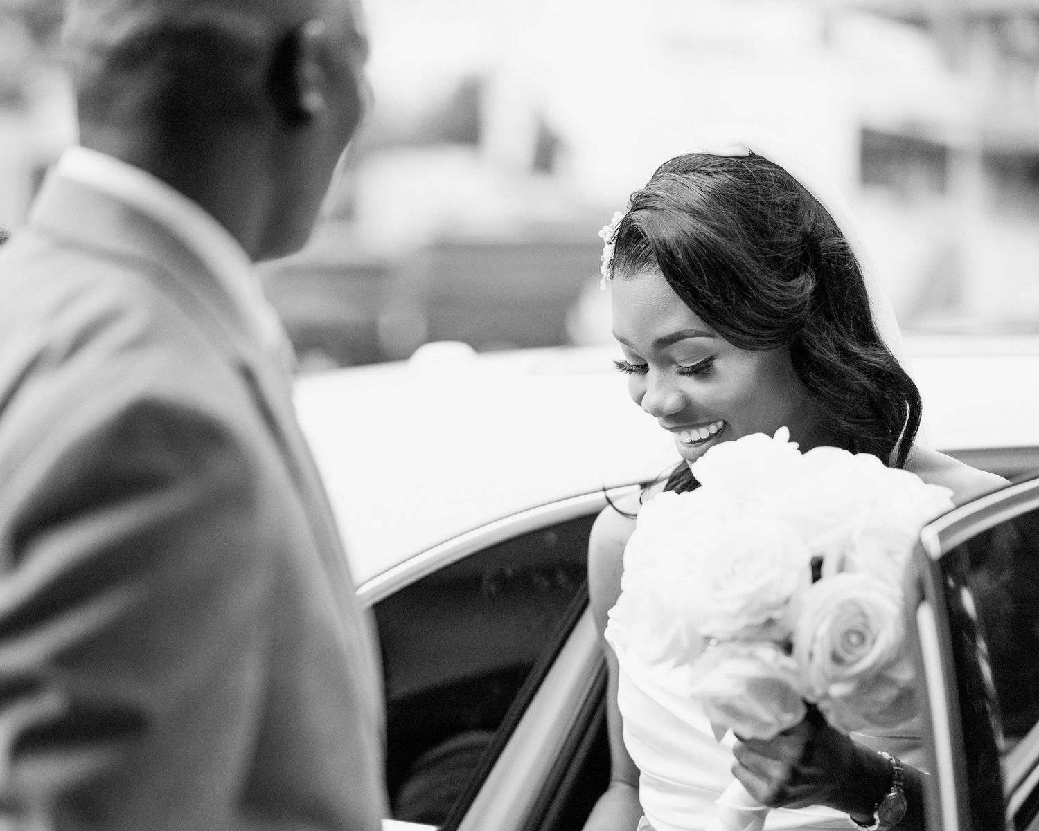 Black and white photo of bride in wedding dress holding white bouquet stepping out of car.