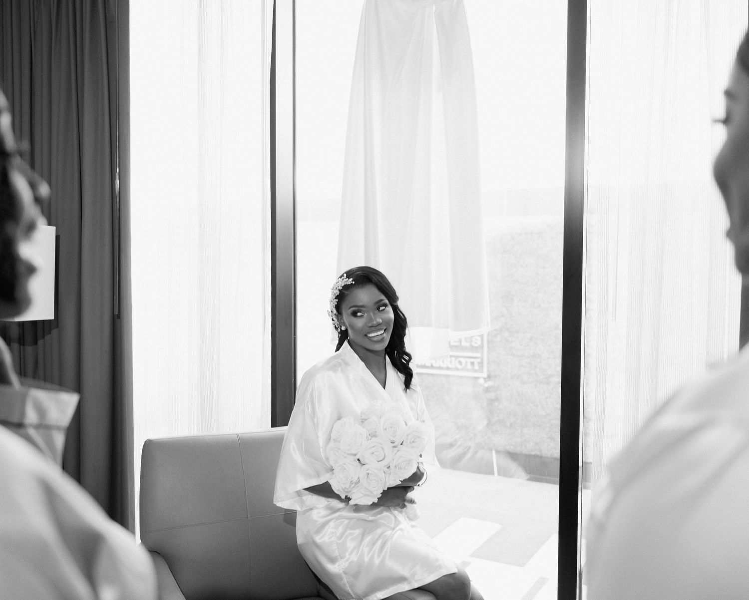 Black and white photo of bride in white robe holding bouquet while sitting during wedding preparations.