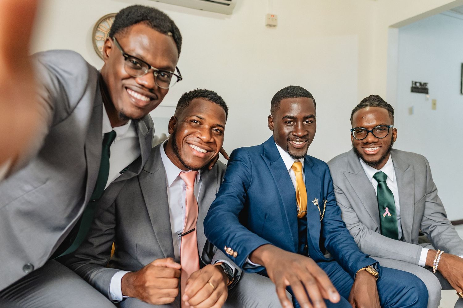 Four well-dressed Black men in suits smiling and taking a selfie together at a wedding celebration.