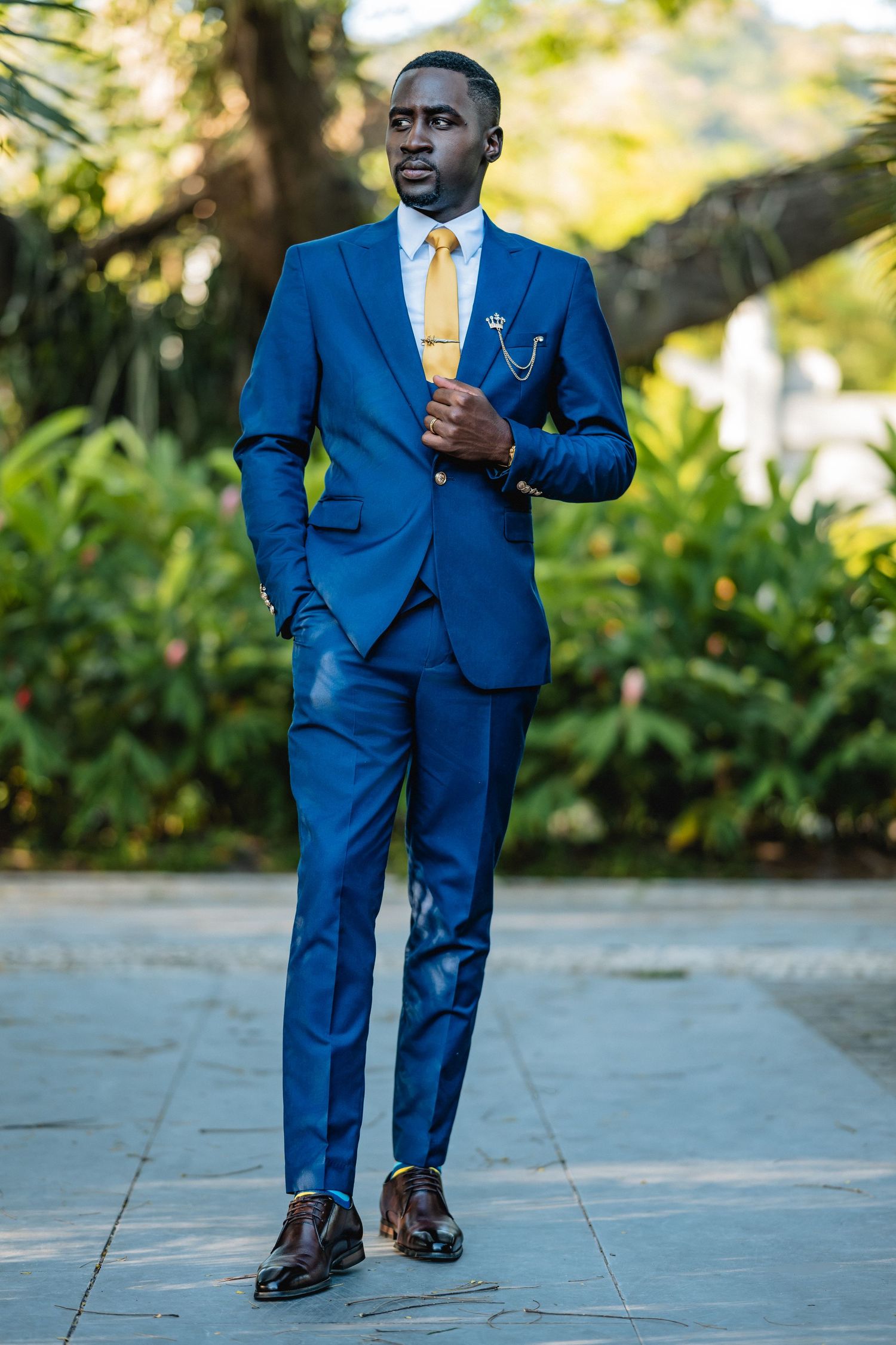 A well-dressed Black man in a blue suit with yellow tie poses outdoors among lush green trees.