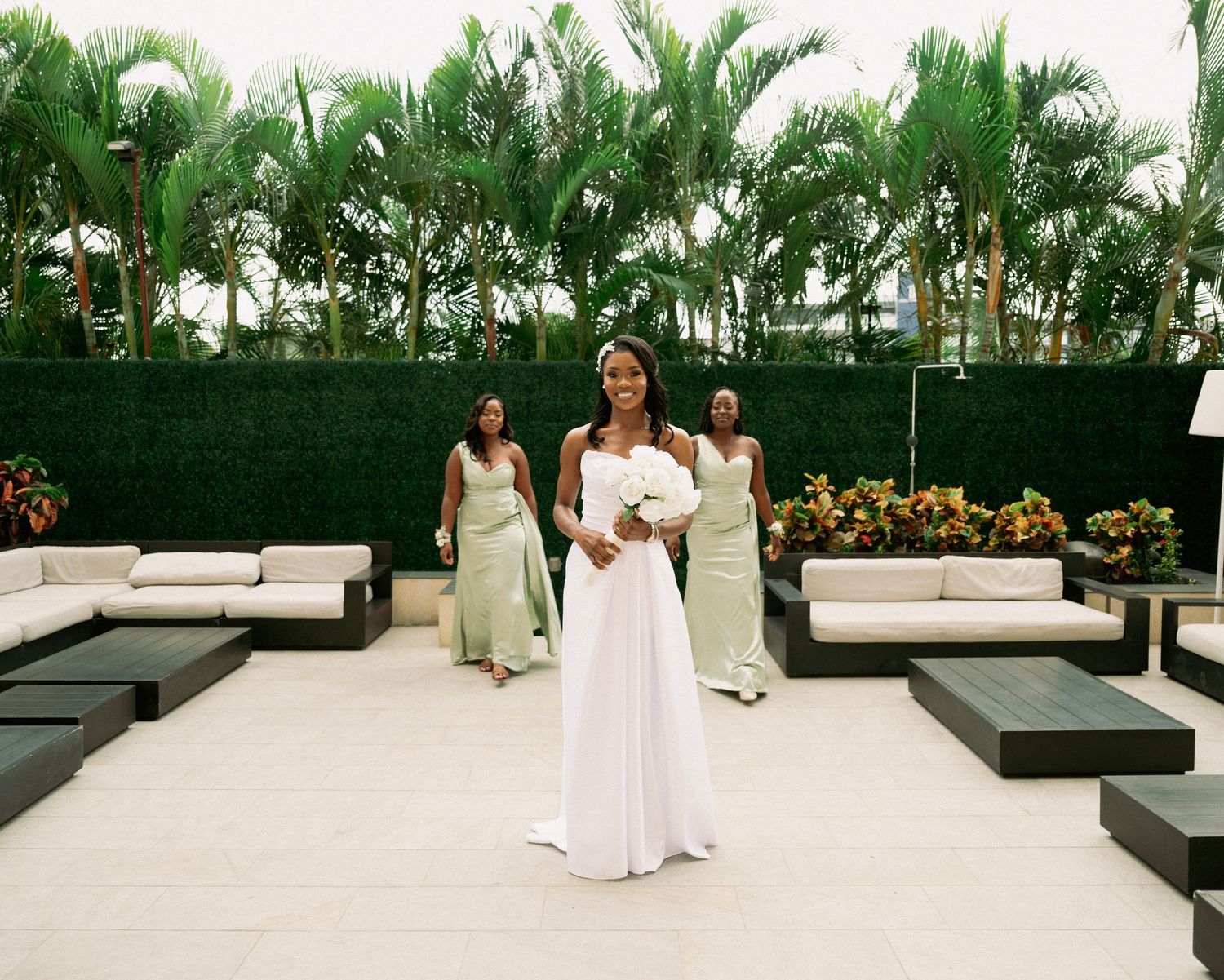 Bride in white gown stands before bridesmaids in sage green dresses on an outdoor patio with palm trees.