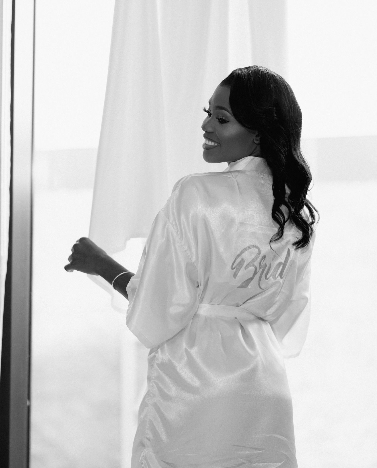Black and white photo of bride in satin Bride robe standing by sheer curtained window.