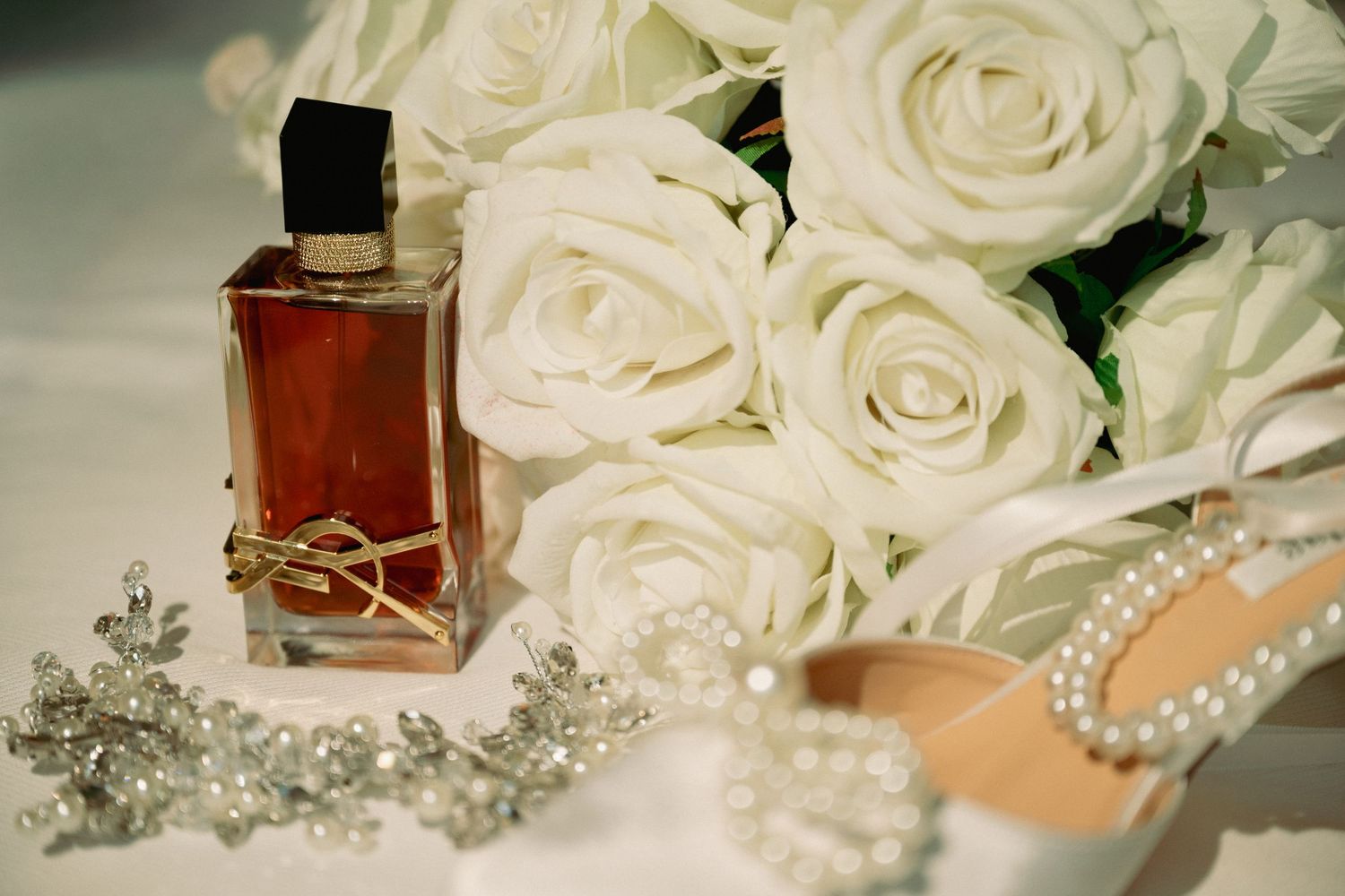 Elegant wedding flat lay with YSL perfume, white roses bouquet, pearl heels, and crystal hair accessories.