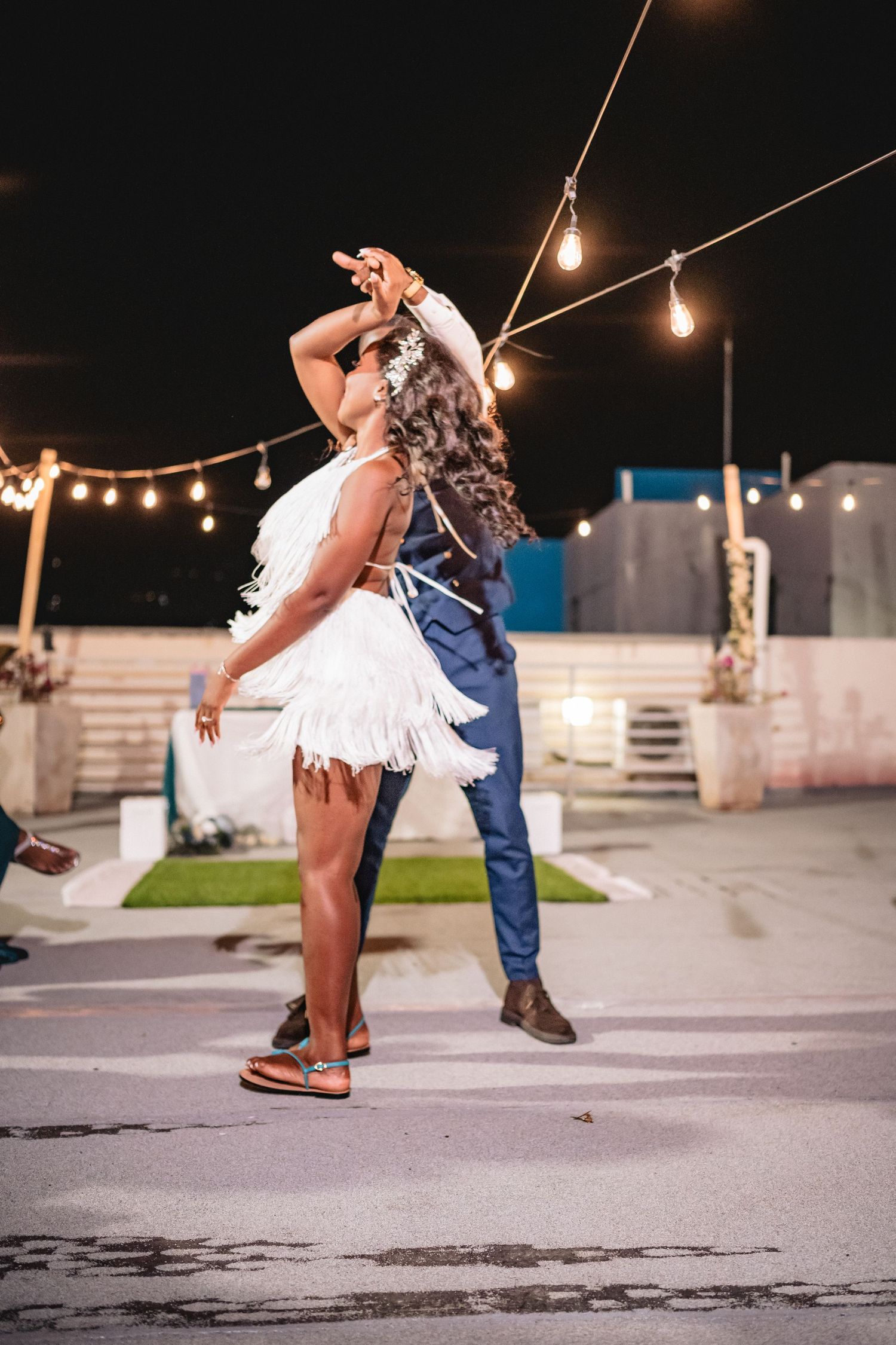 Bride in white fringe dress dancing energetically with groom under string lights at outdoor night wedding reception.