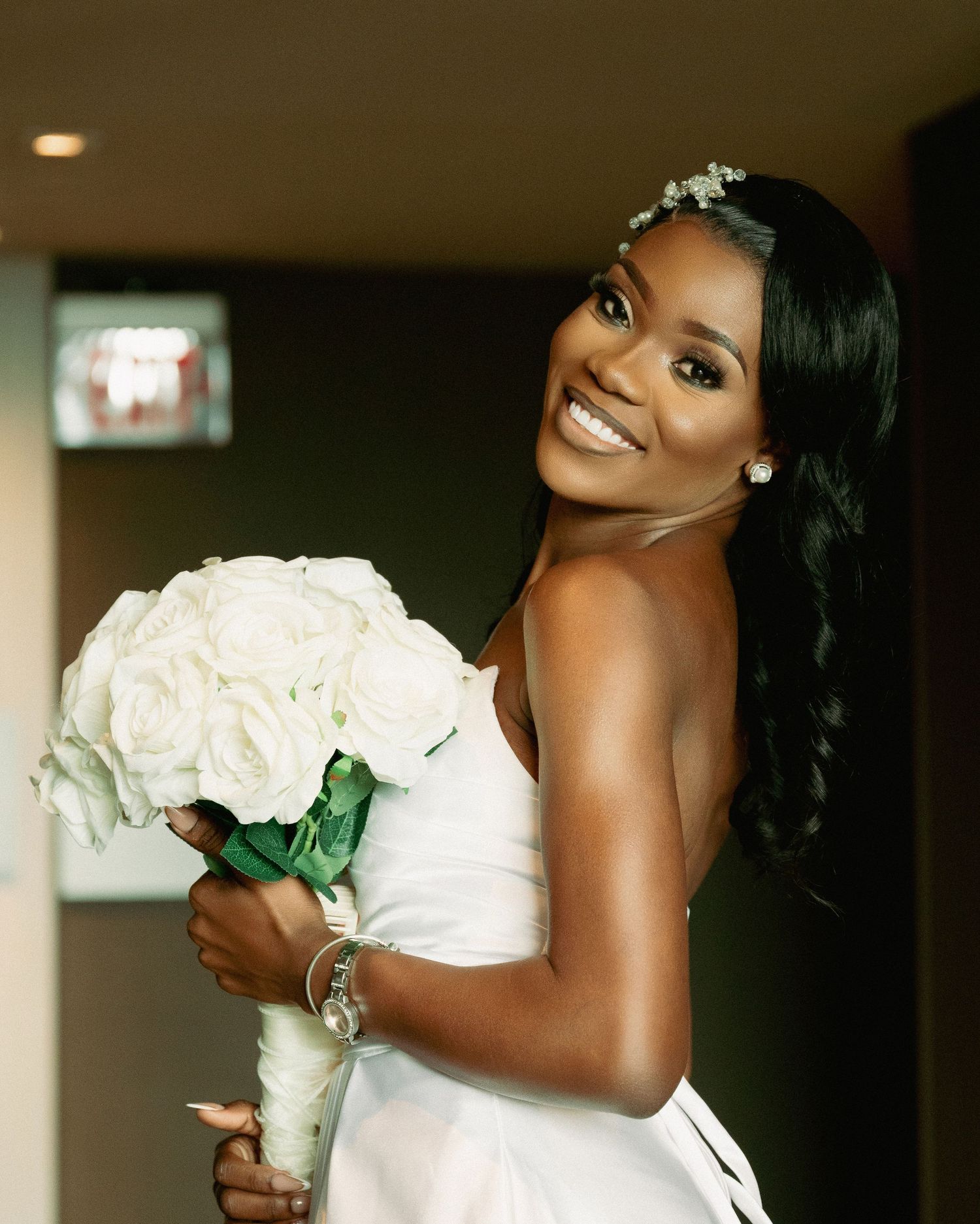 Elegant bride with white rose bouquet poses in indoor venue with dramatic lighting.