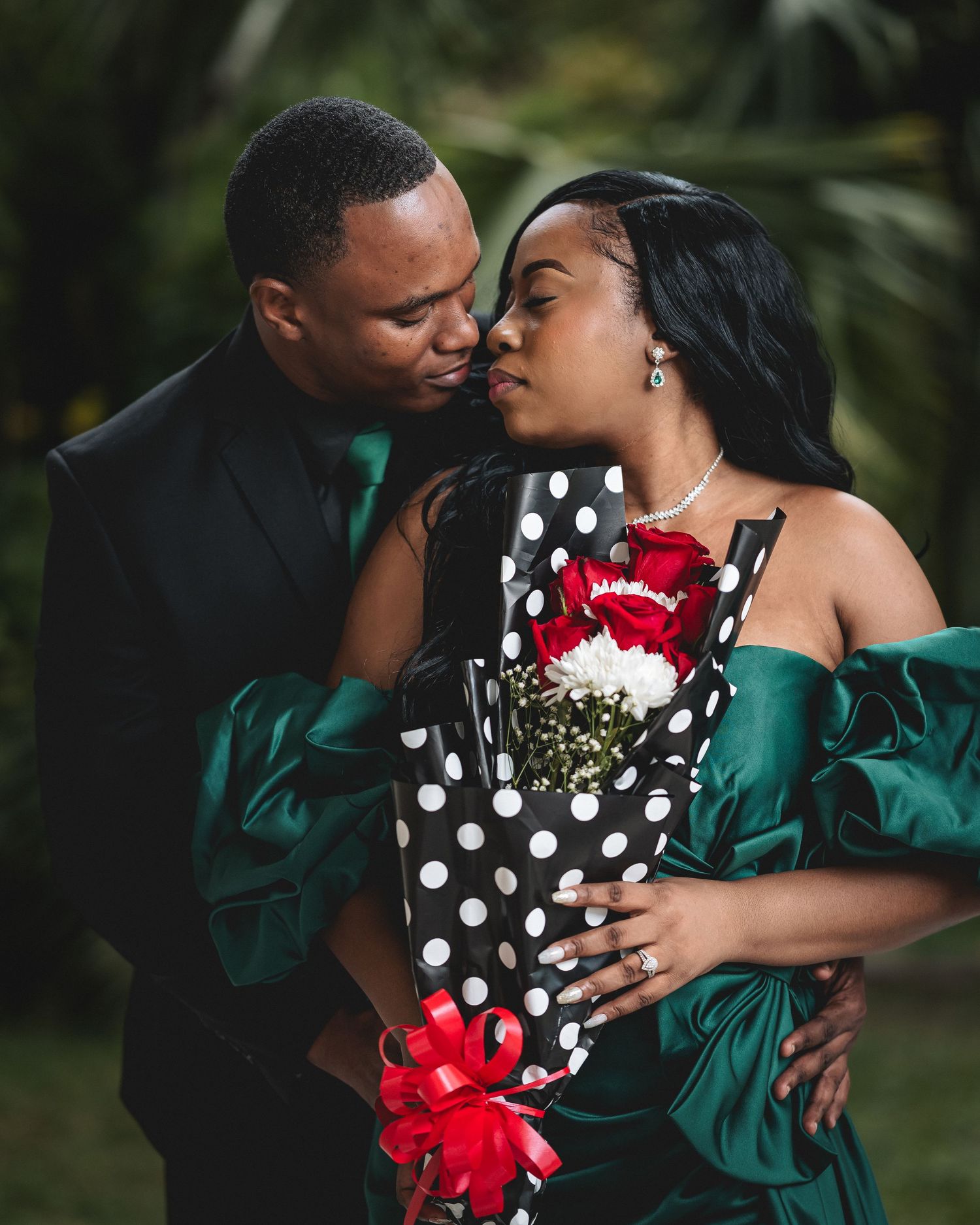Couple in elegant green outfits sharing a romantic moment, woman holding red roses bouquet with polka dot wrapping.