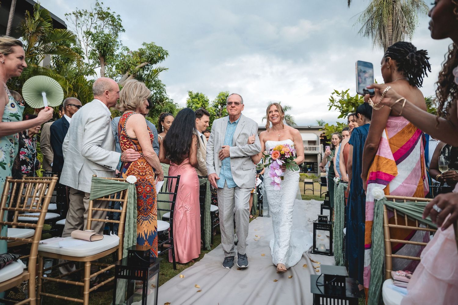 Bride walks down outdoor aisle with escort at tropical wedding ceremony, guests standing alongside decorated chairs.
