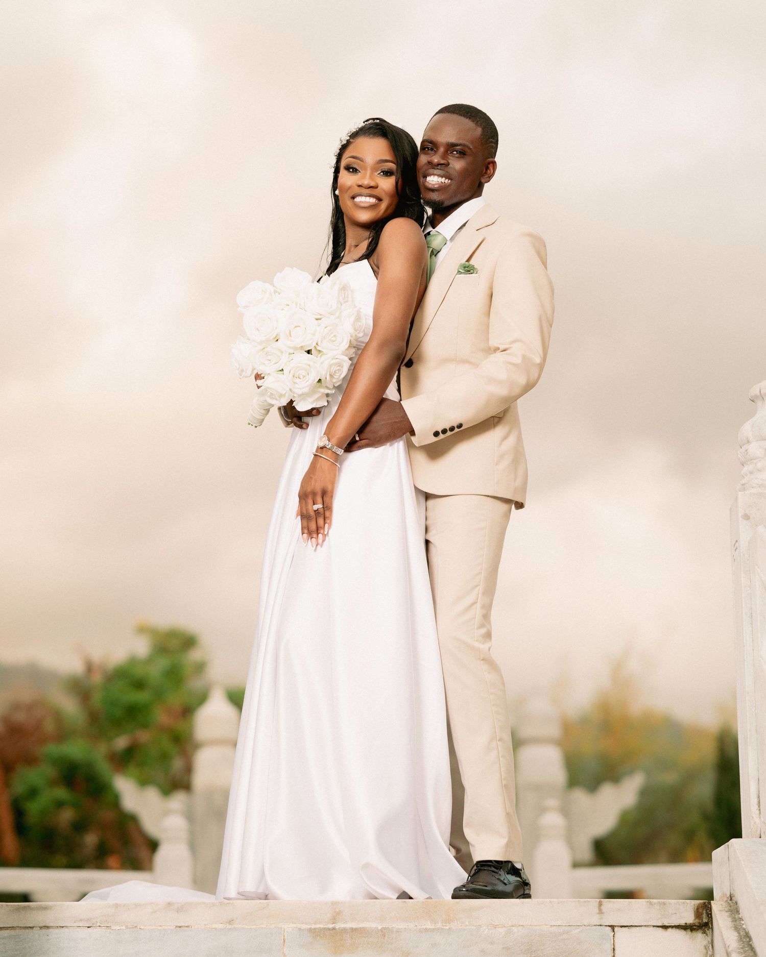 Happy couple on wedding day, bride in white gown holding bouquet, groom in beige suit outdoors.