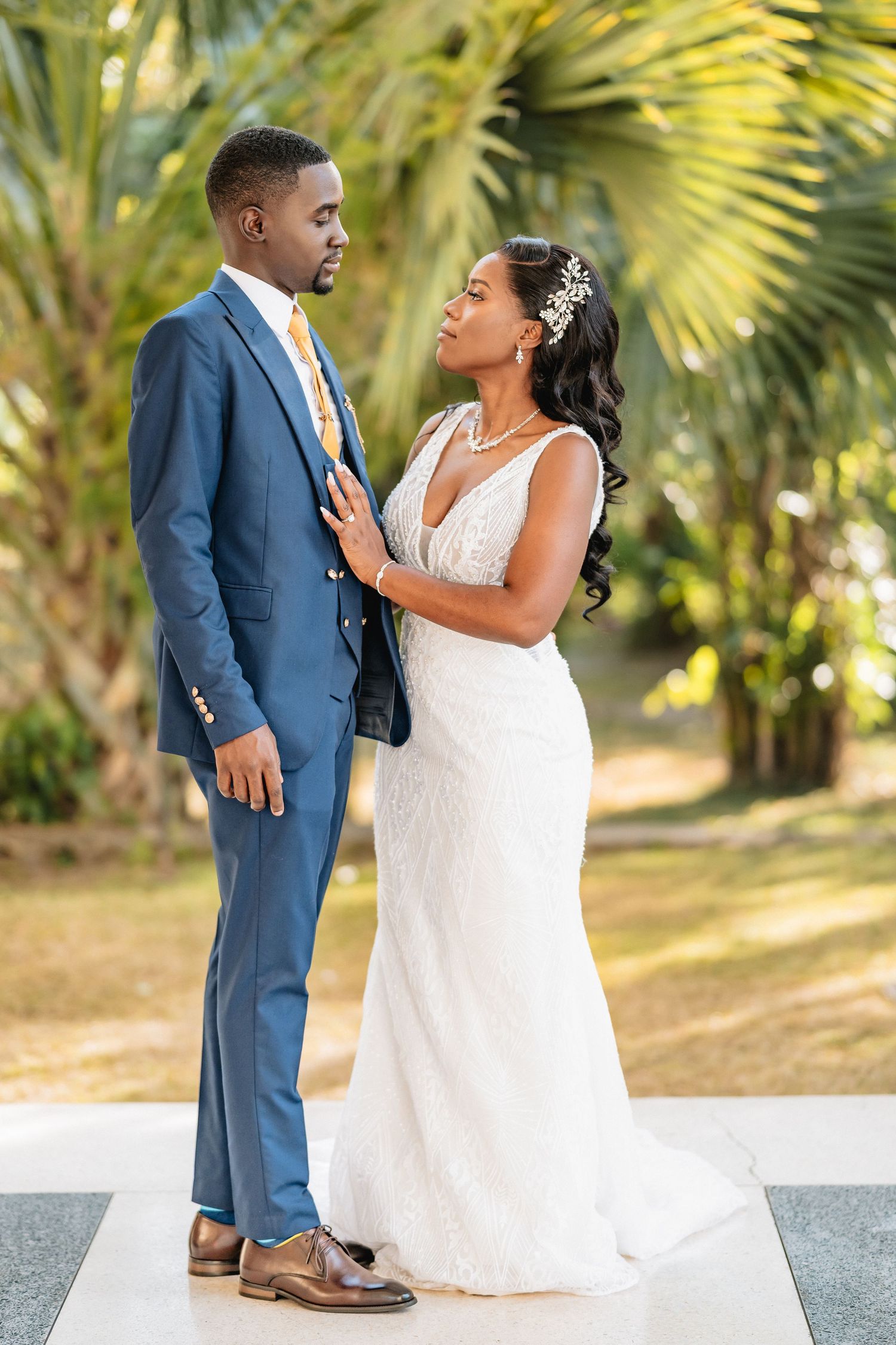 Elegant bride and groom in blue suit share a romantic moment outdoors with tropical palm trees backdrop.