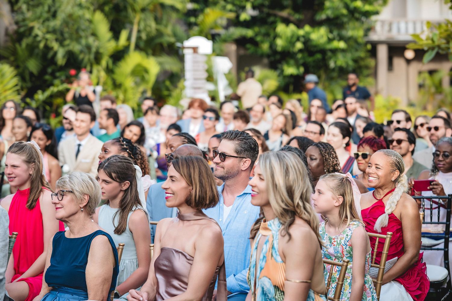 Smiling wedding guests gathered outdoors at a sunny garden ceremony, dressed in colorful summer attire.