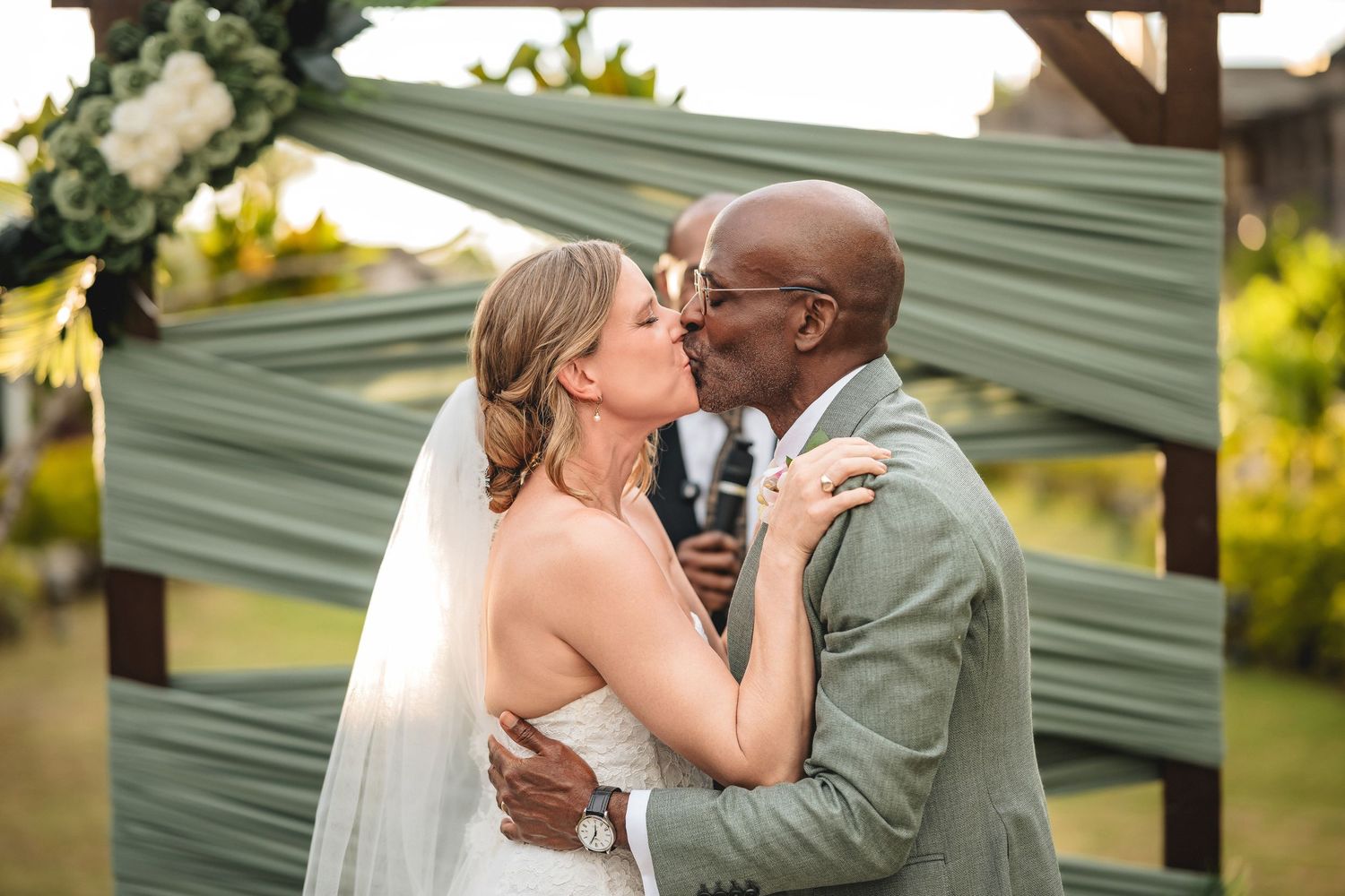 Interracial couple shares first kiss at outdoor wedding ceremony beneath sage green draped arch.