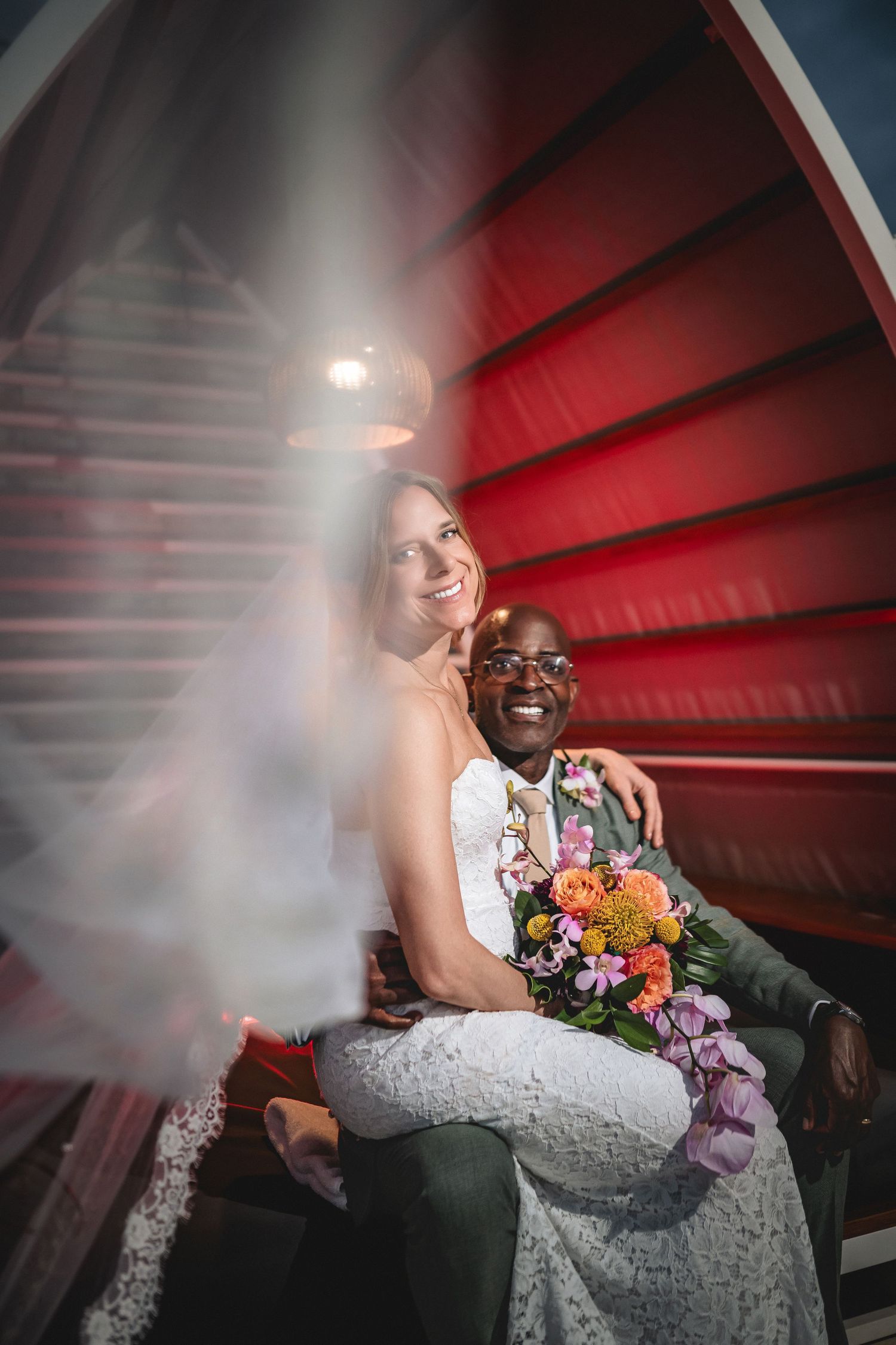 Interracial couple in wedding attire sitting together, bride in white lace dress holding colorful bouquet, groom smiling, red backdrop.