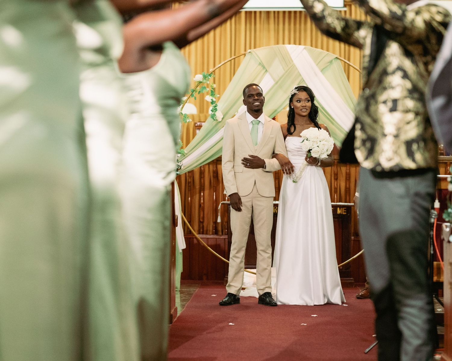 Couple stands at church altar beneath gold circular arch with green and white draped decorations.