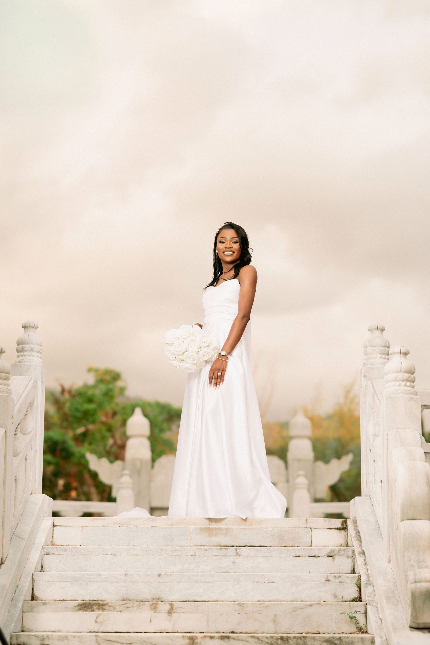 Elegant bride in white gown holds bouquet atop white stone staircase under dramatic cloudy sky.
