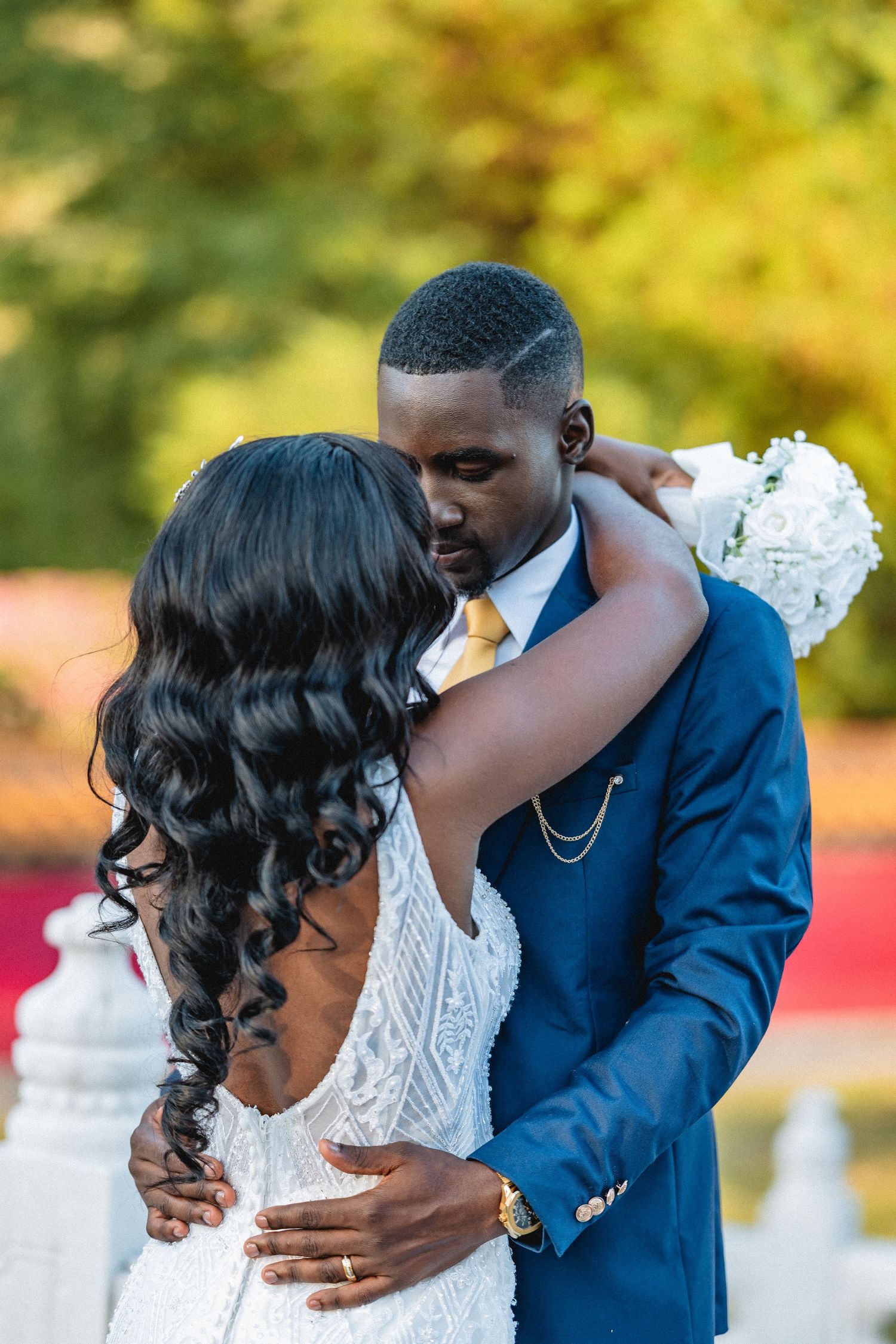 Bride and groom share romantic embrace outdoors, she holds white bouquet in elegant wedding attire.