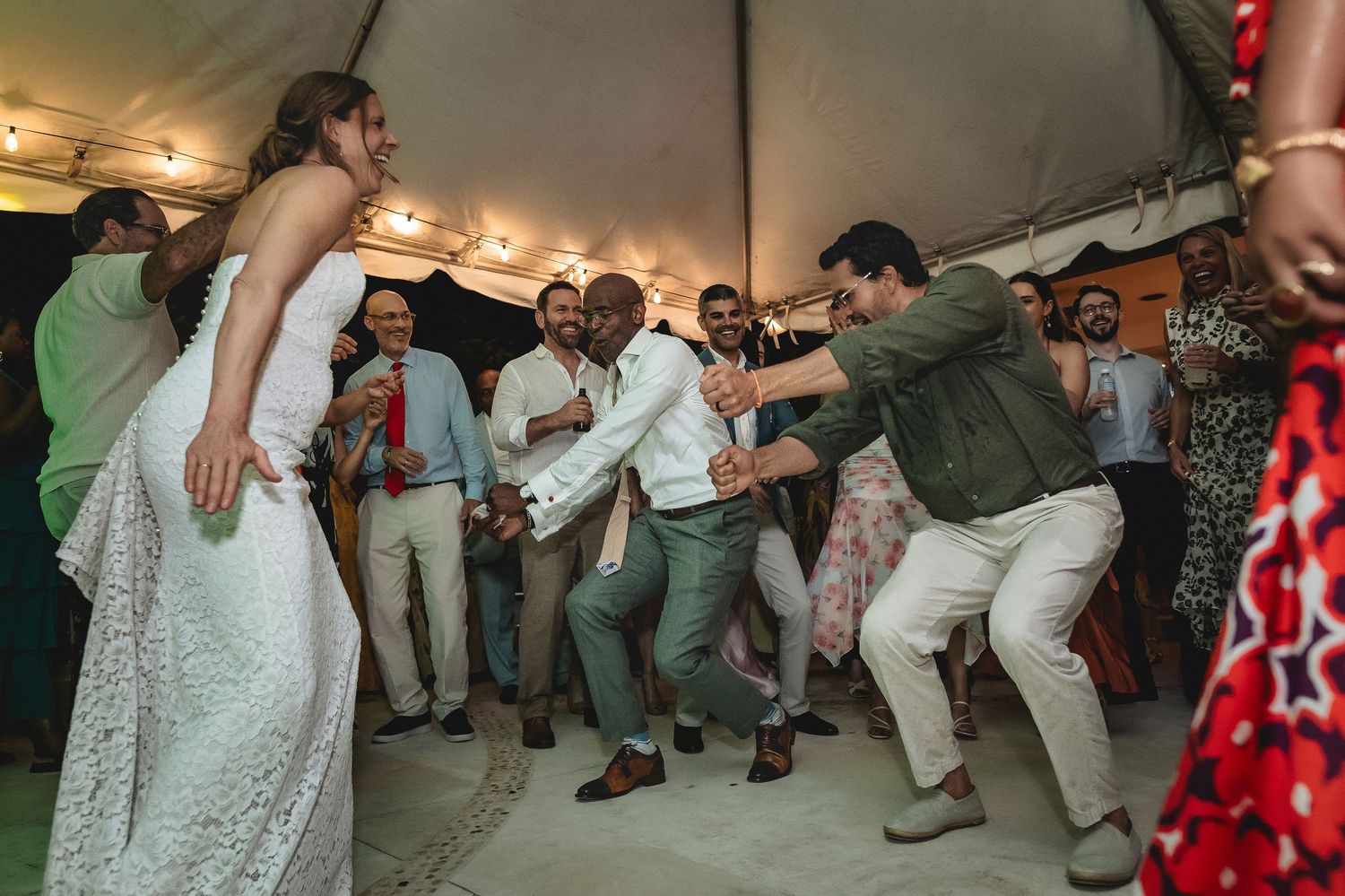 Wedding guests dancing energetically at a festive outdoor reception under a tent with string lights.