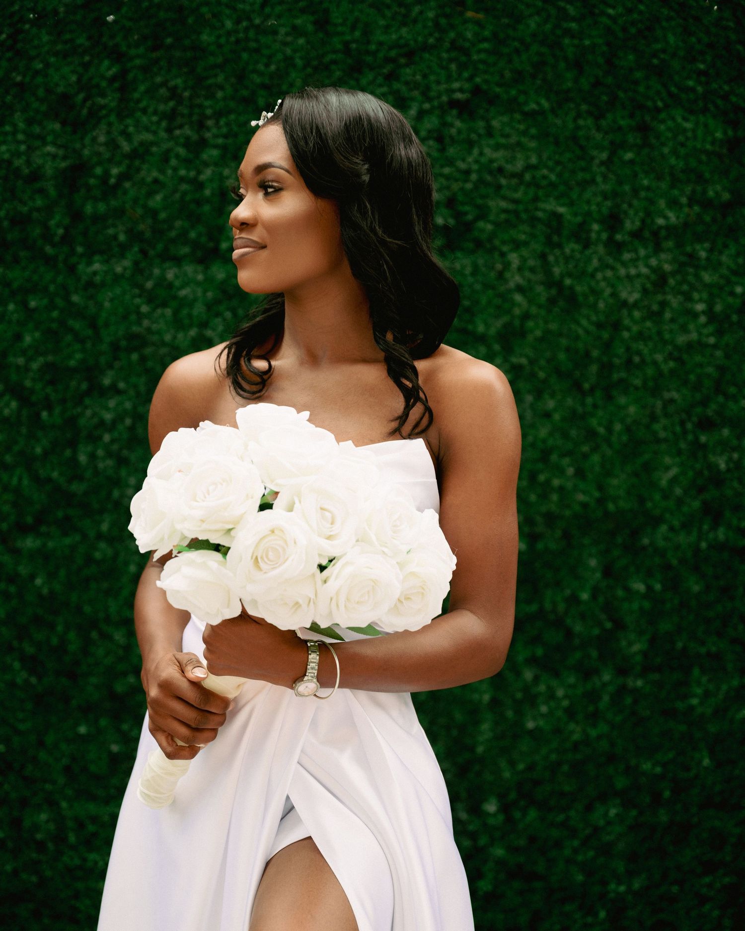 Beautiful Black bride in strapless white gown holding lush white rose bouquet in front of green hedge wall.