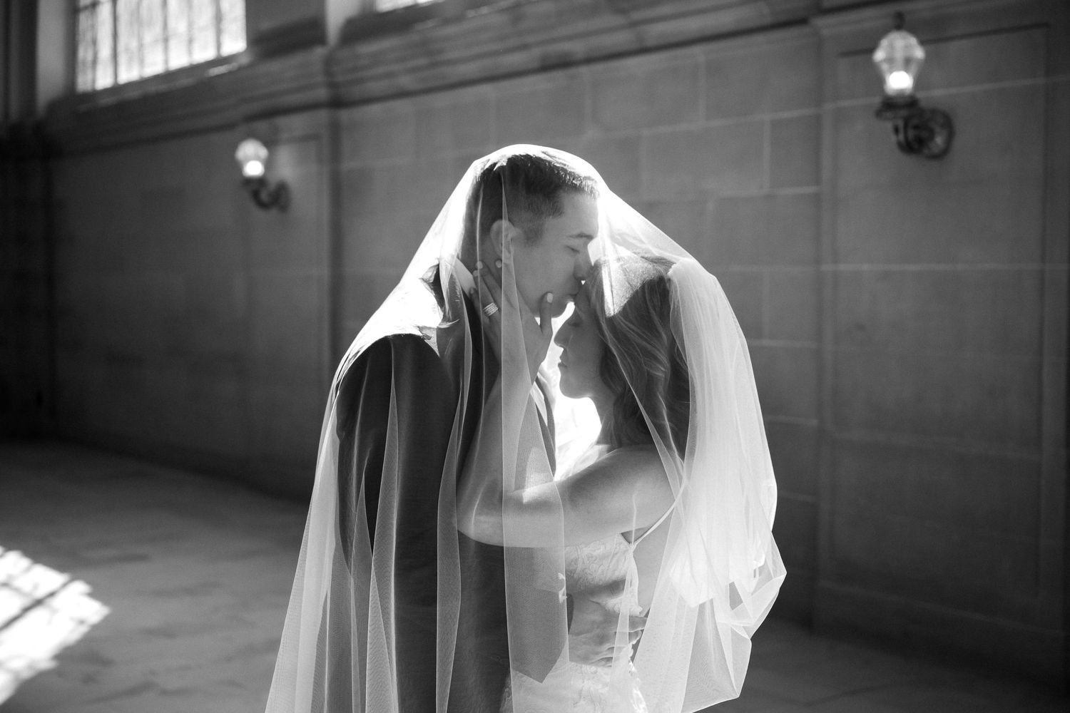 Black and white photo of bride and groom sharing a romantic kiss under a flowing wedding veil in an elegant venue.