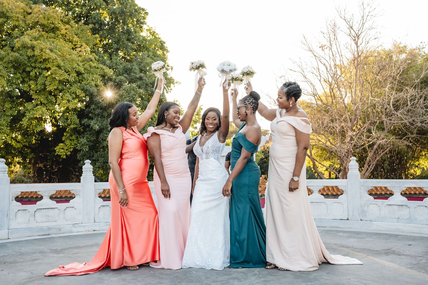Four bridesmaids in coral and teal gowns celebrate with a bride in a white wedding dress outdoors.