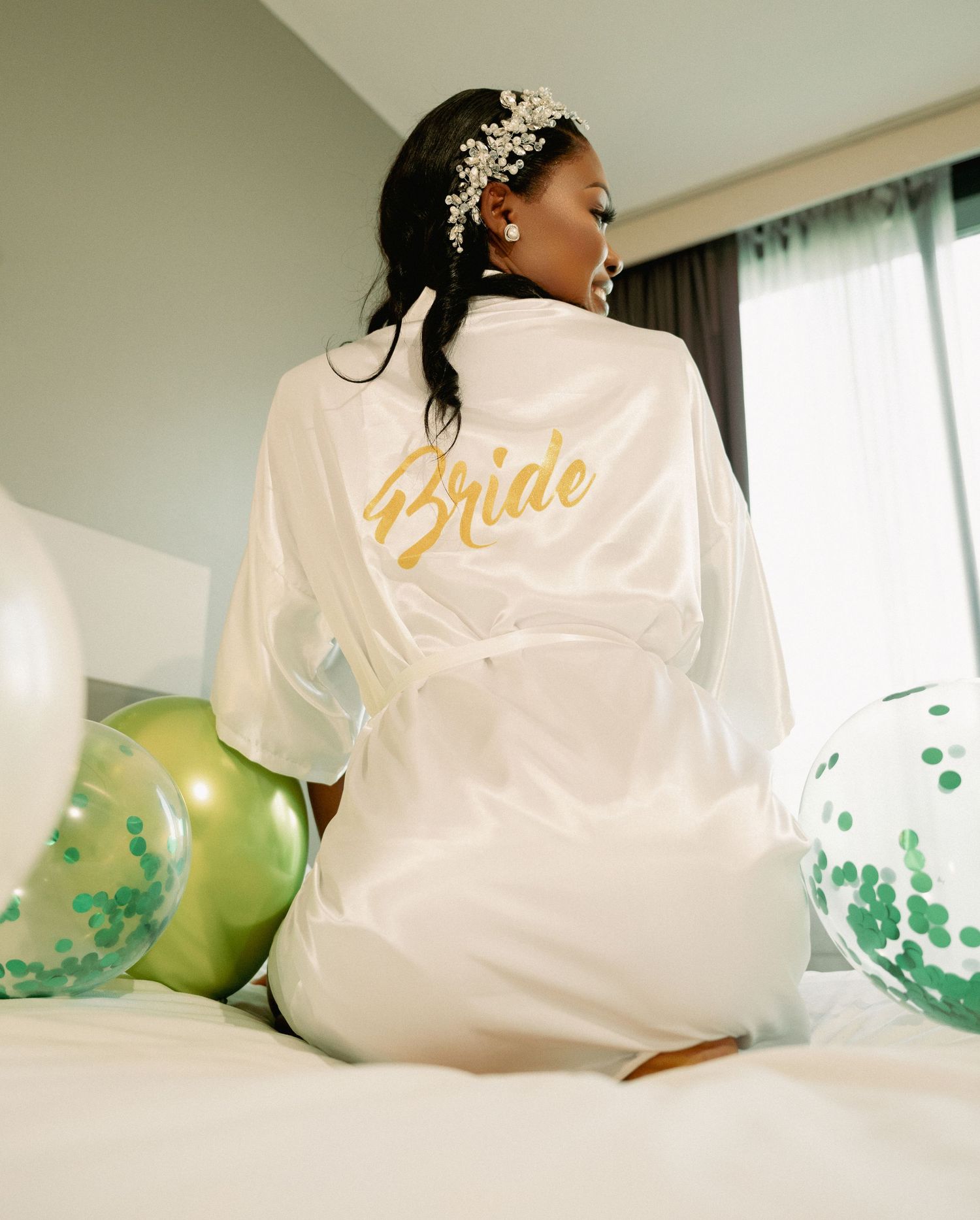A bride in a white satin robe with gold Bride text sits among balloons.