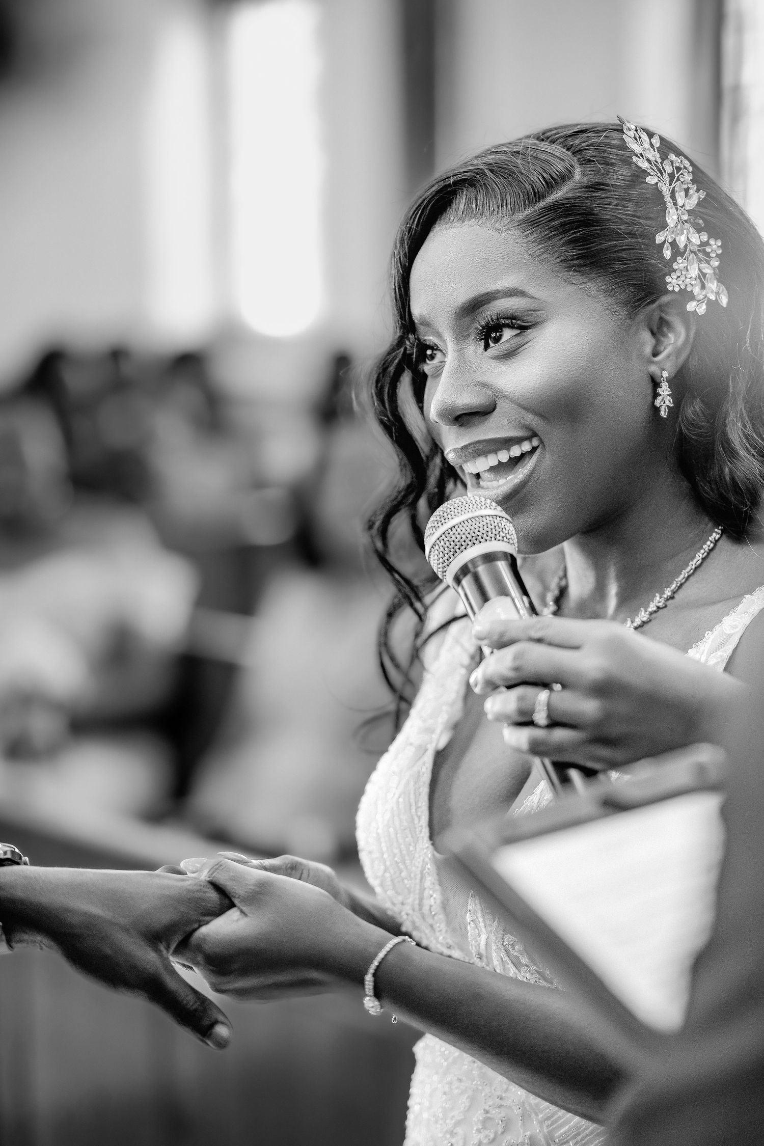 Smiling Black bride in white lace dress holds microphone during wedding ceremony speech, wearing hair accessories and jewelry.