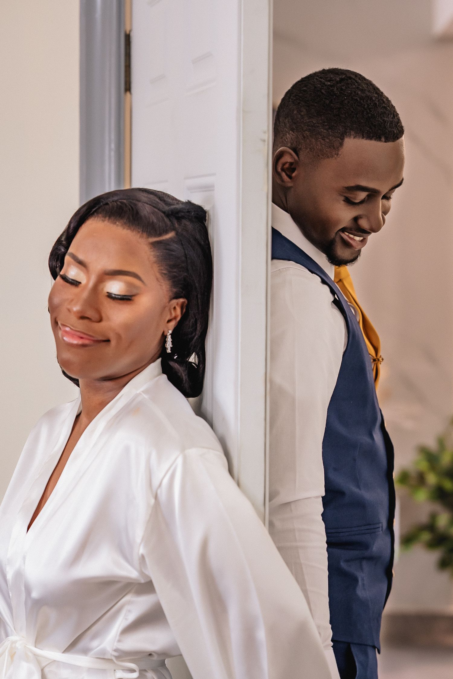 A bride in white robe and groom in blue suit lean back-to-back against a door, smiling on their wedding day.