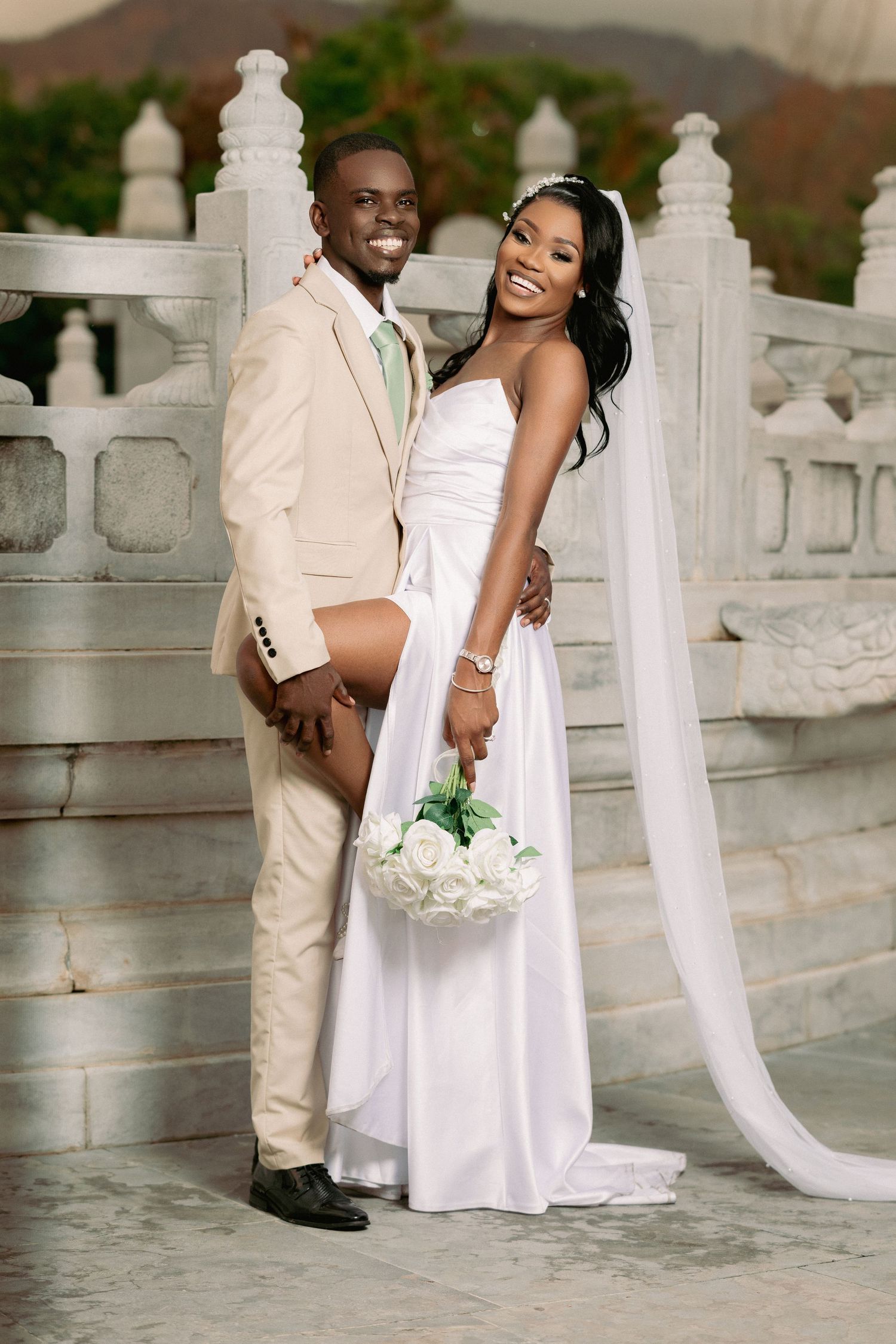 Happy Black couple in wedding attire pose on marble steps, bride in white gown with veil holding white roses.
