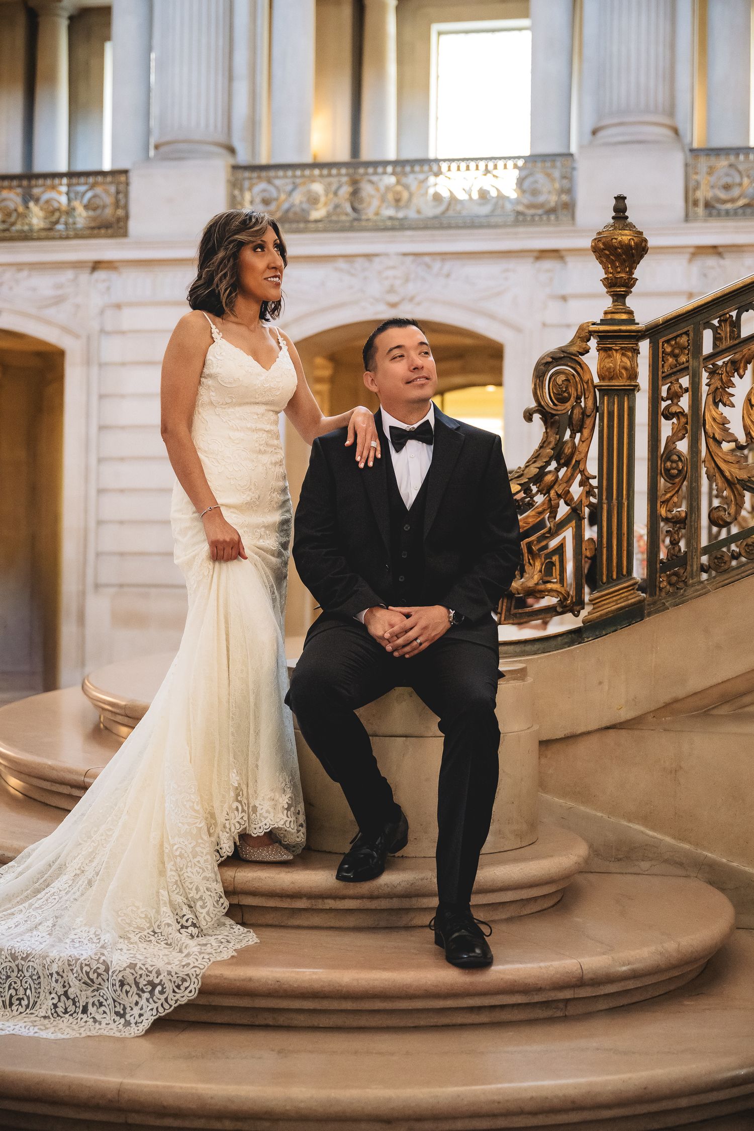 Bride in lace gown and groom in tuxedo pose on ornate staircase at San Francisco City Hall wedding.