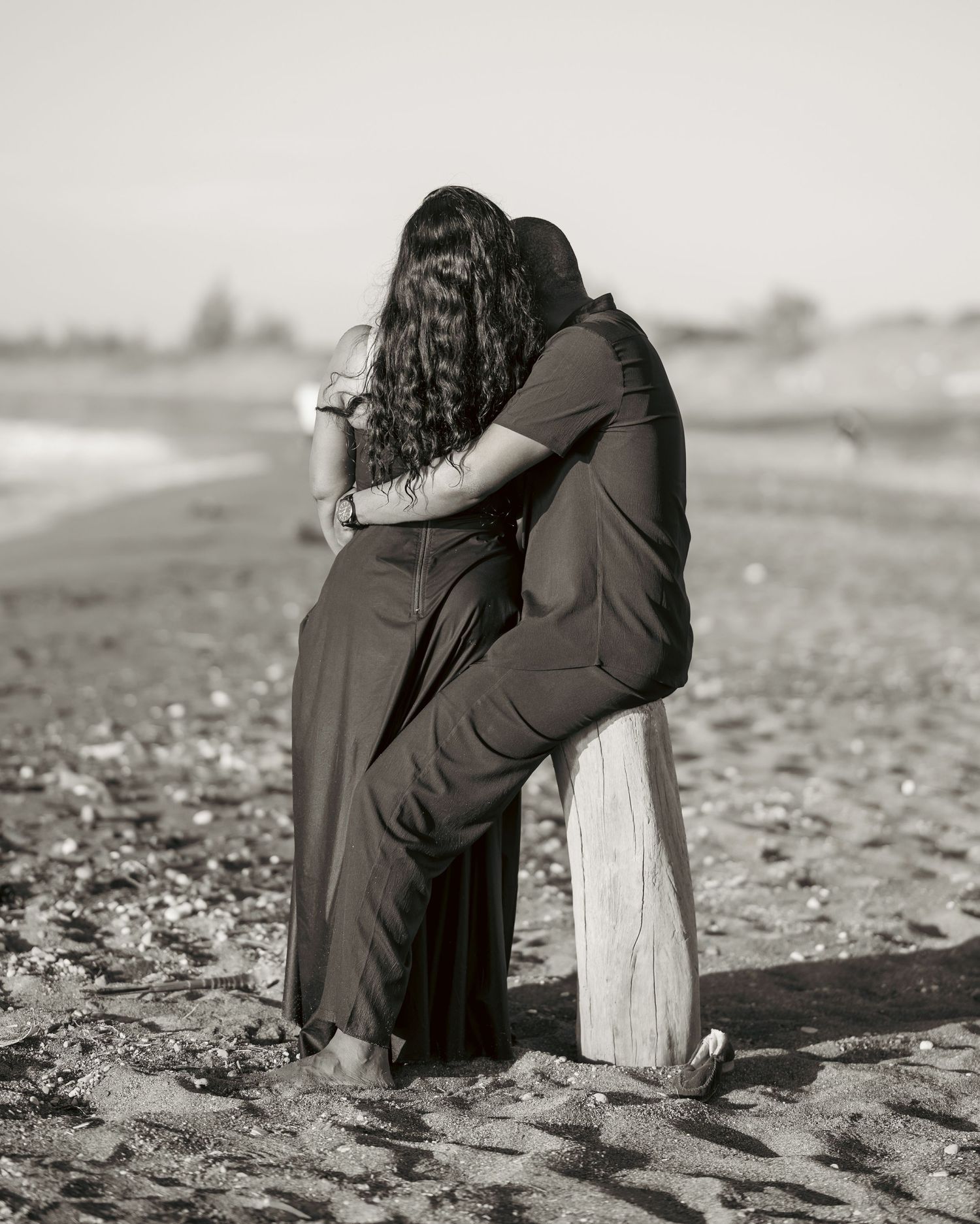 Black and white photo of a couple embracing on a sandy beach, woman in long dress leaning into mans arms.