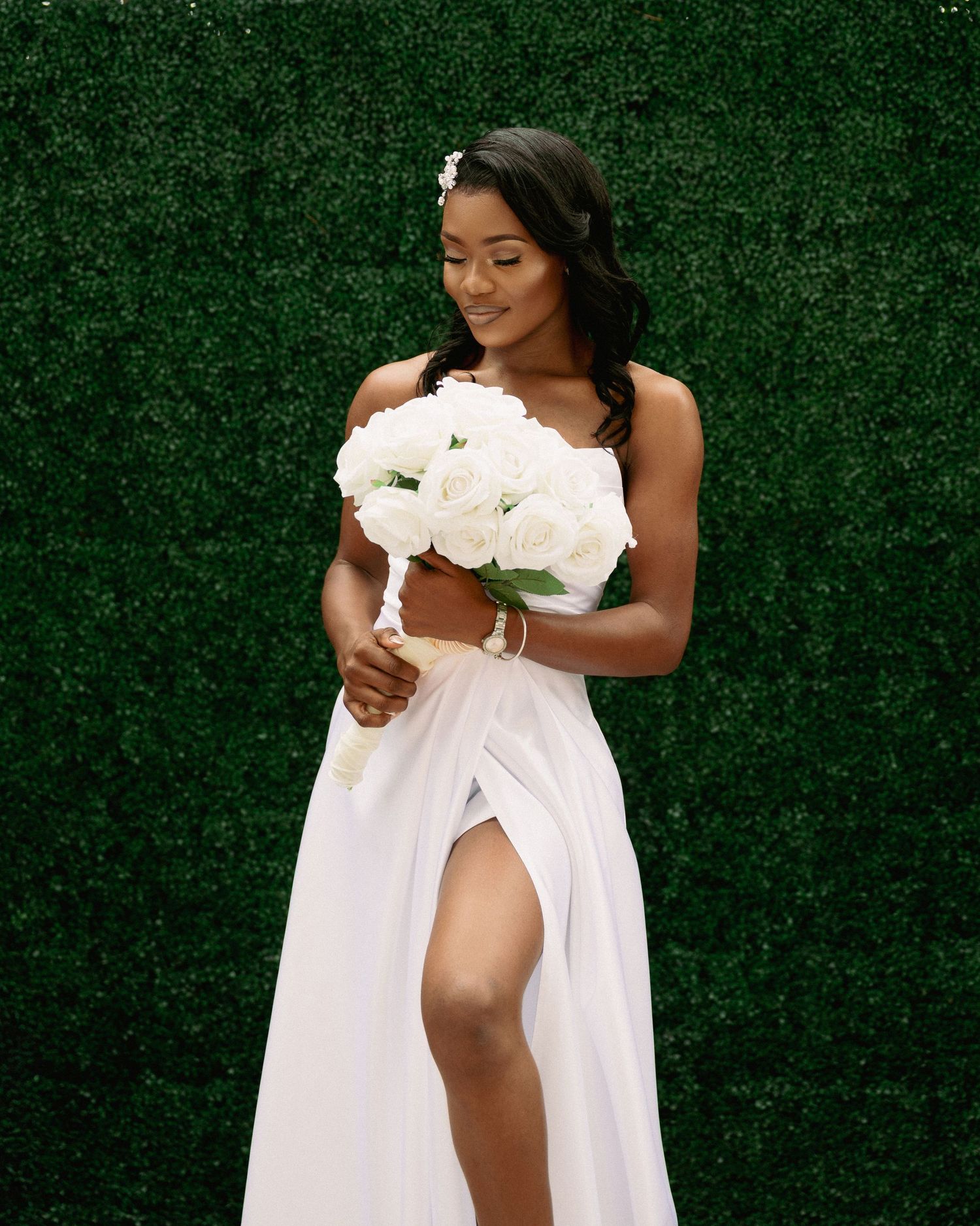 Elegant Black bride in a white dress with a slit holding white roses in front of a green hedge wall.