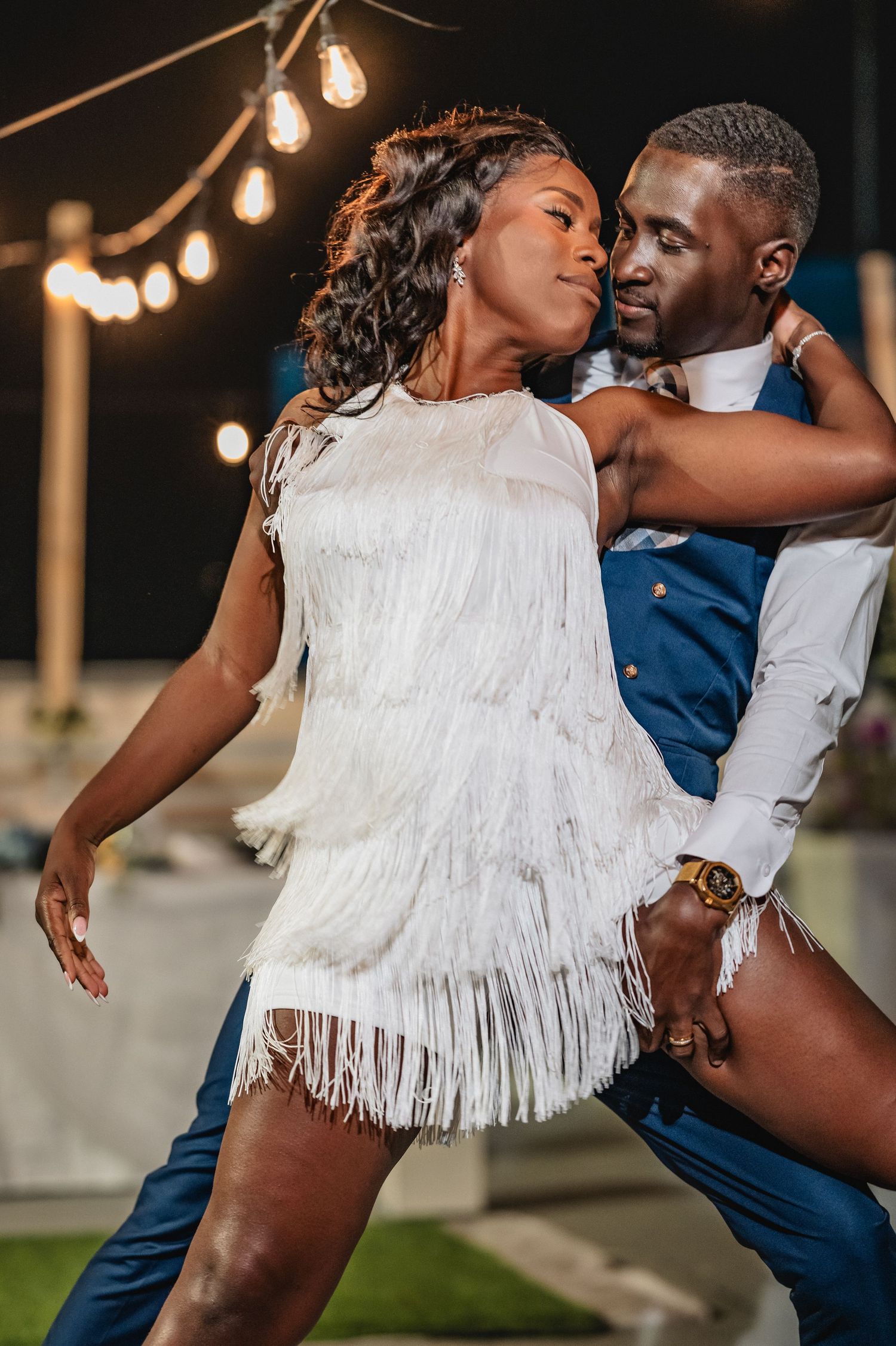 Bride in white fringe dress and groom in blue suit share energetic dance at outdoor reception.