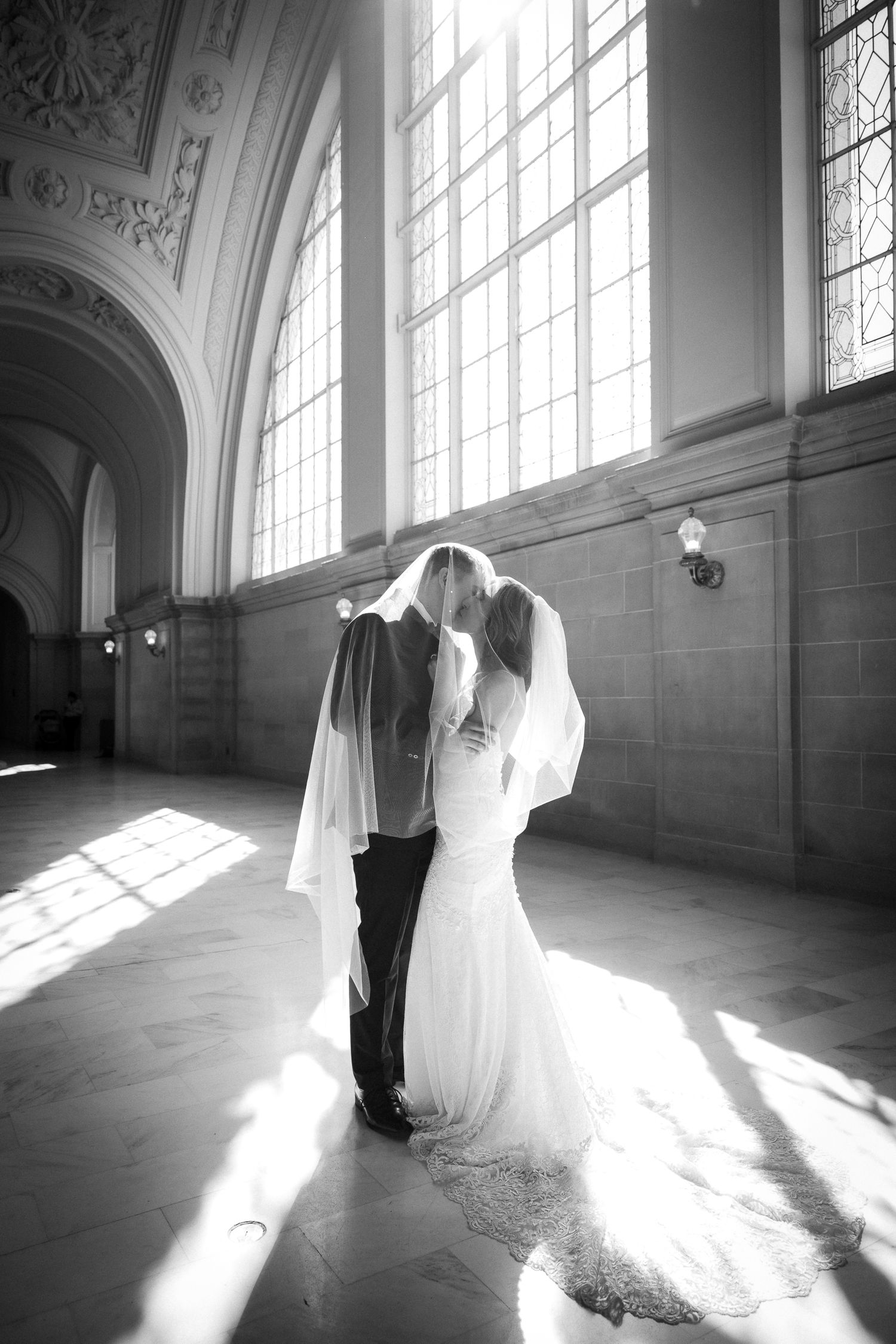 Black and white photo of bride and groom sharing a kiss under veil in sunlit grand hall.