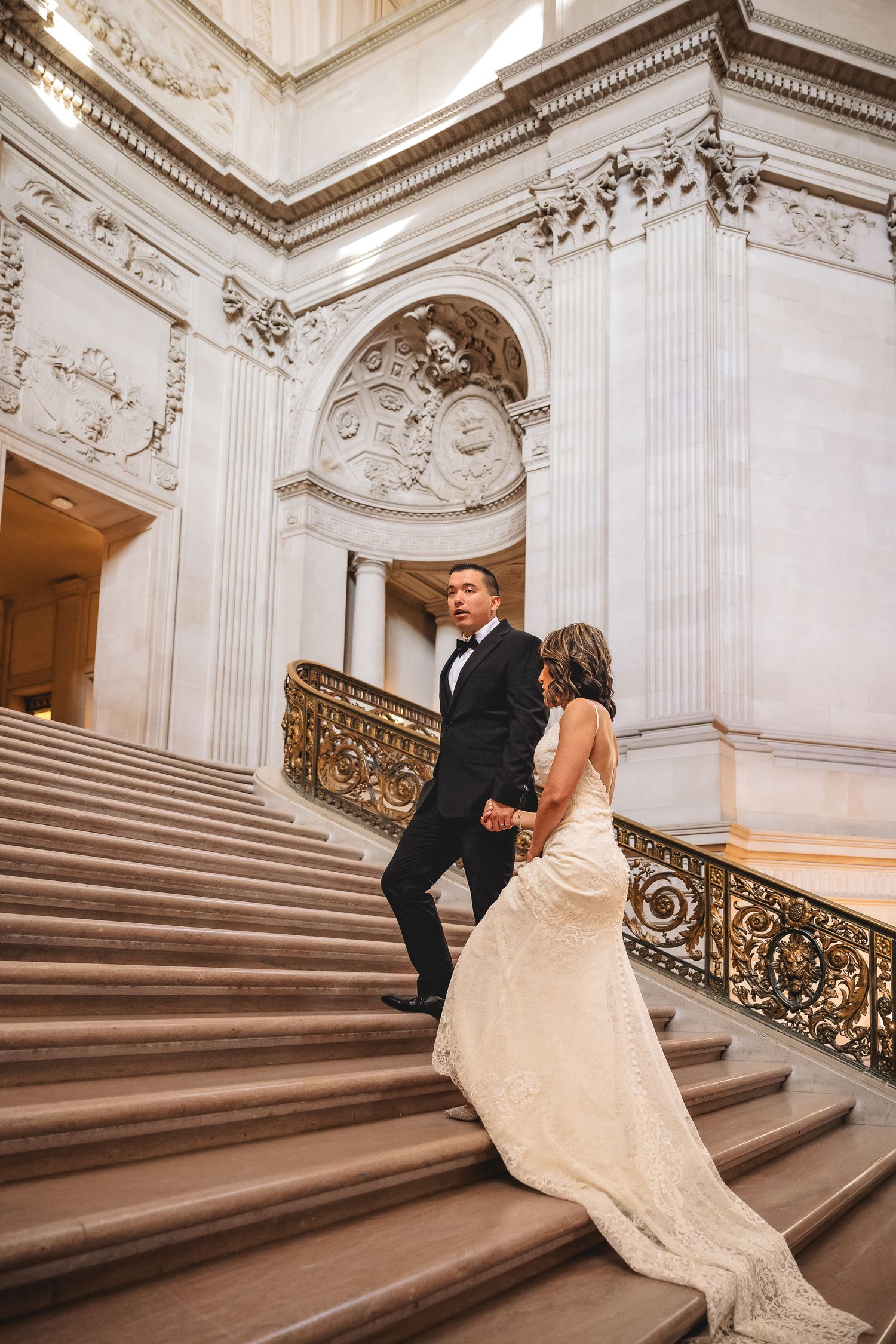 Bride and groom walking up grand staircase in elegant white marble venue, bride in lace gown, groom in black tuxedo.