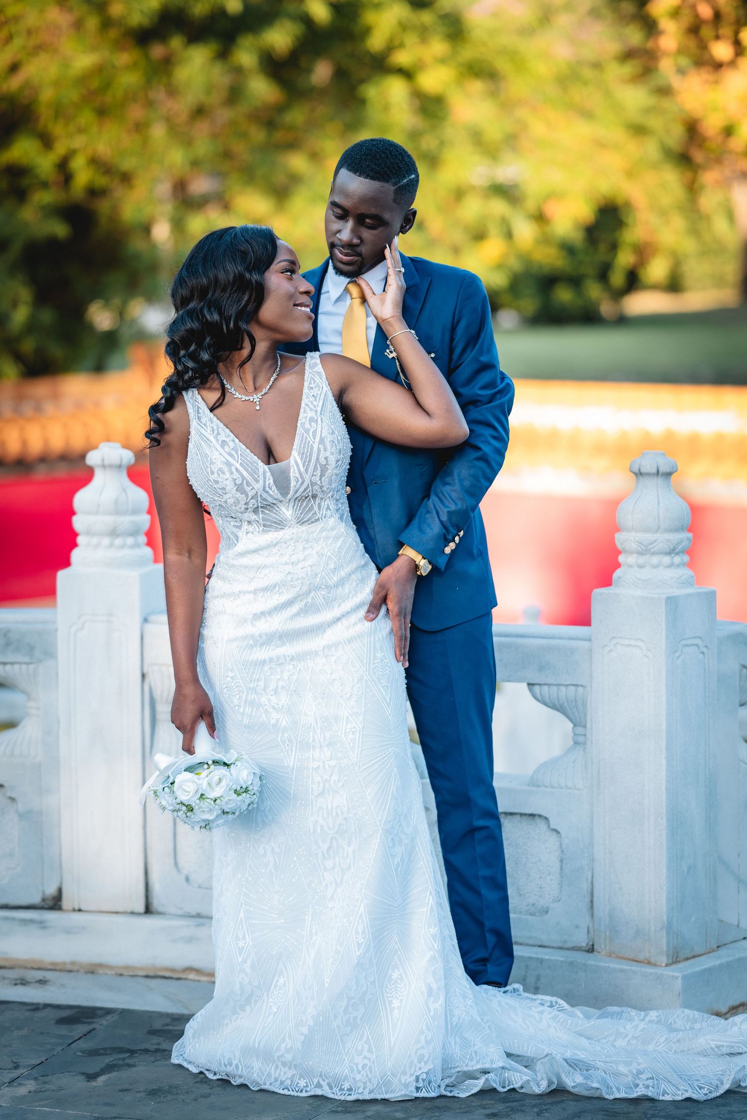 Black bride and groom share romantic moment outdoors, she in white lace gown with bouquet, he in blue suit.