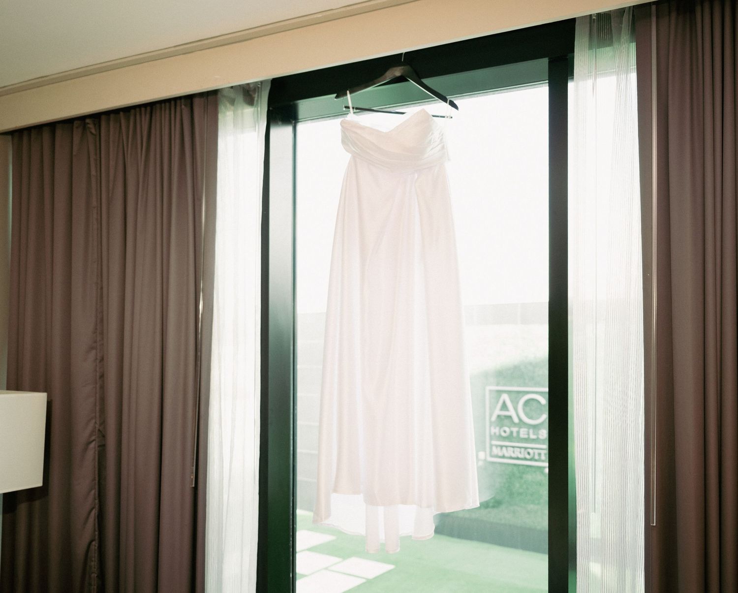 White strapless wedding gown hanging on a hanger in front of a window at AC Hotel.