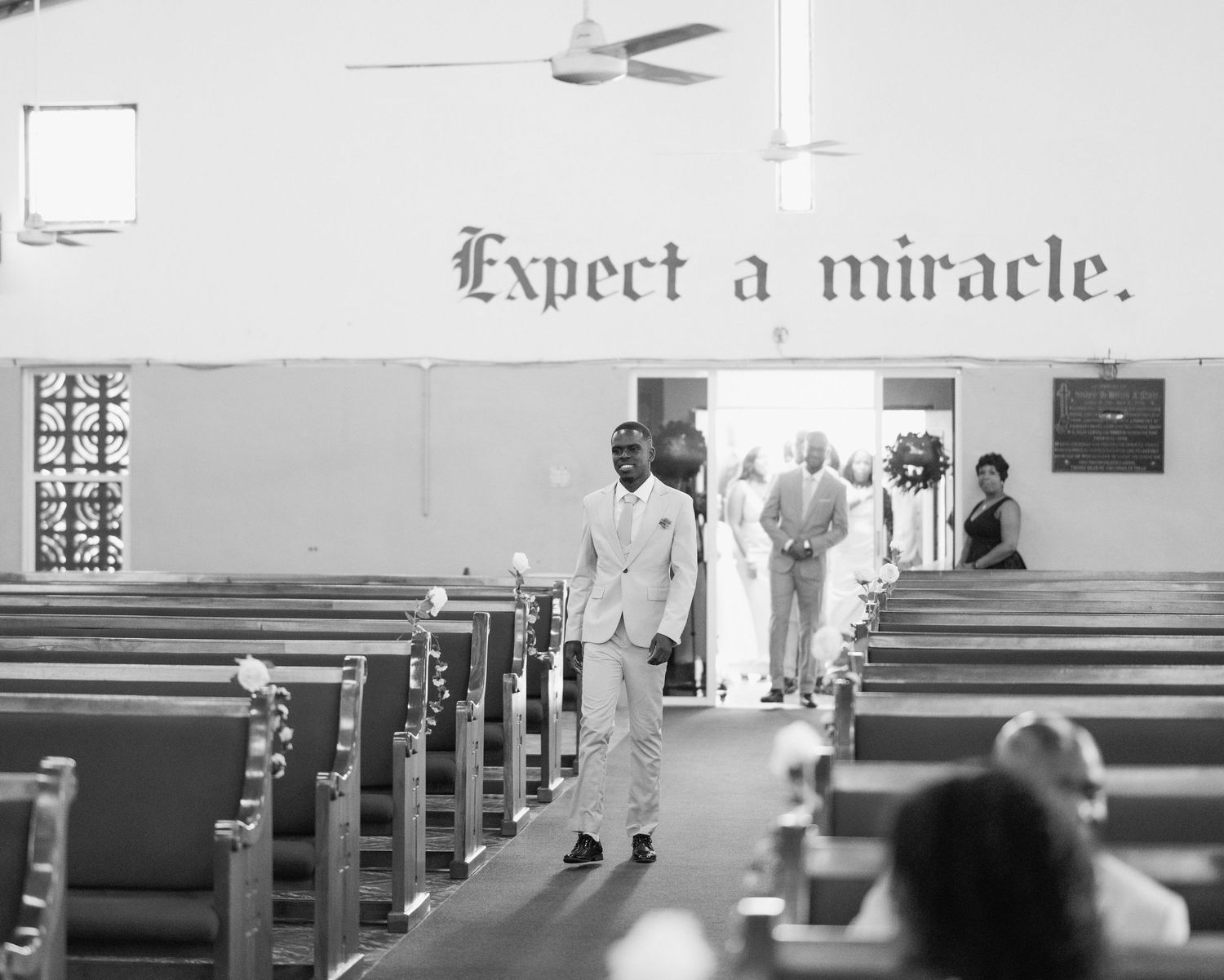 Groom in white suit walks church aisle under Expect a Miracle sign in black and white photo.