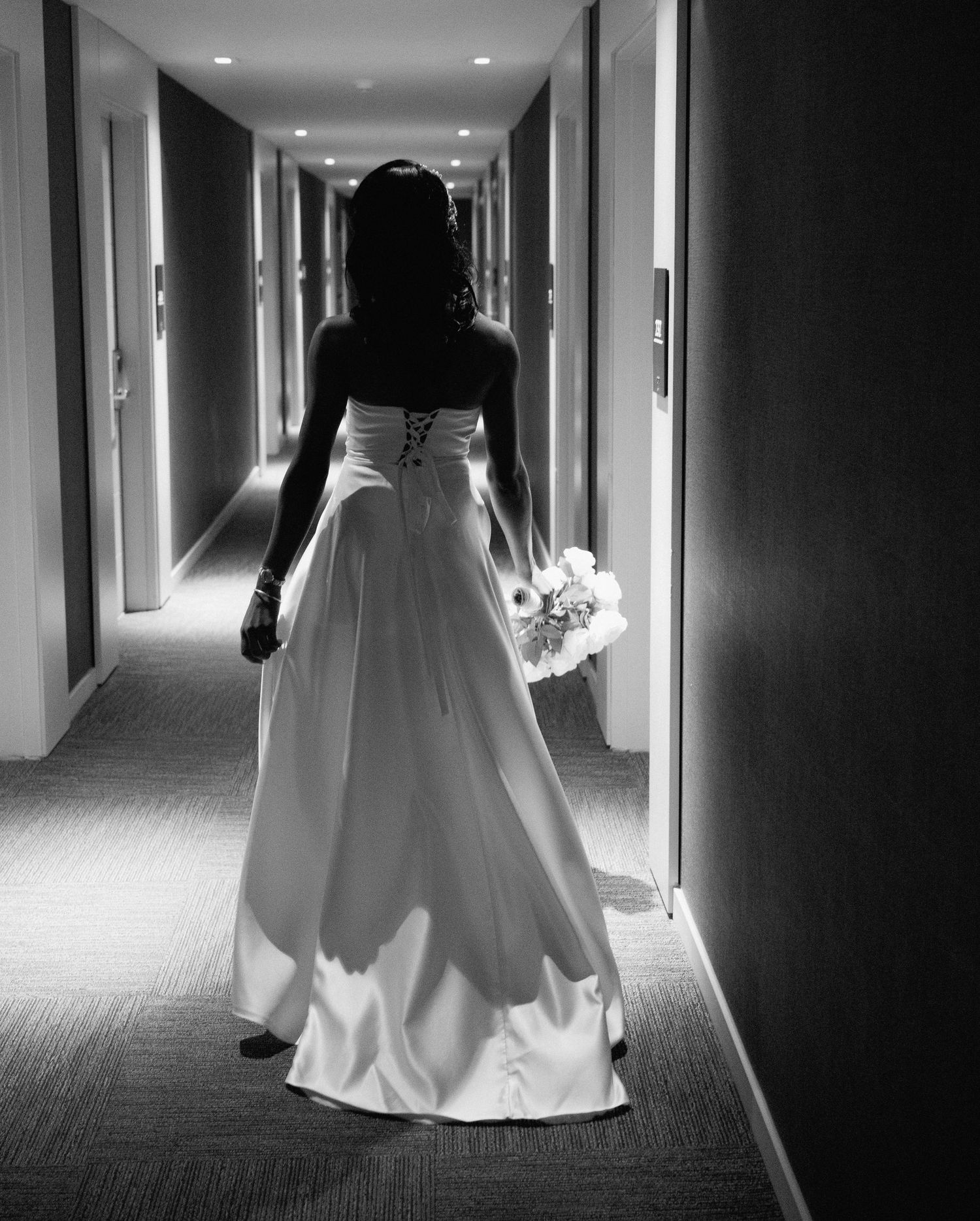 Black and white photo of a bride in a strapless gown walking down a hotel hallway with her bouquet.