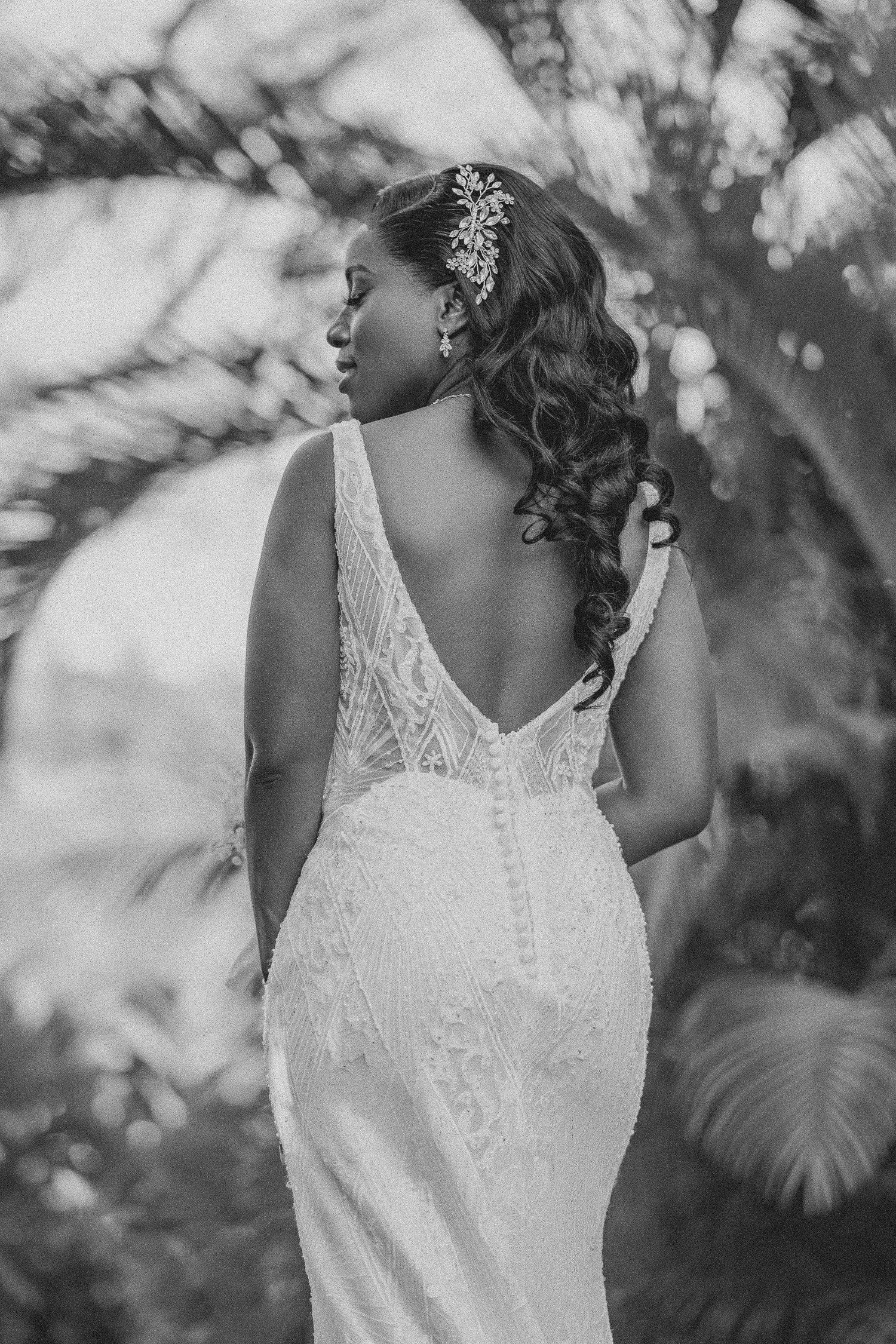 Black and white portrait of bride in backless lace wedding gown with floral hair accessory among palm trees.