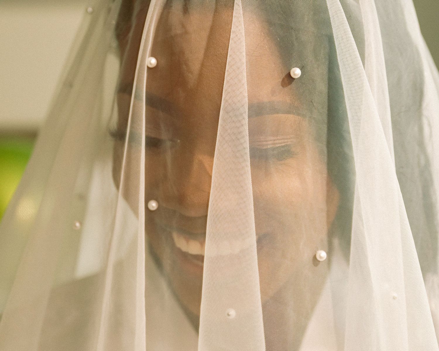 Smiling bride wearing a pearl-adorned wedding veil covering her face on her wedding day.