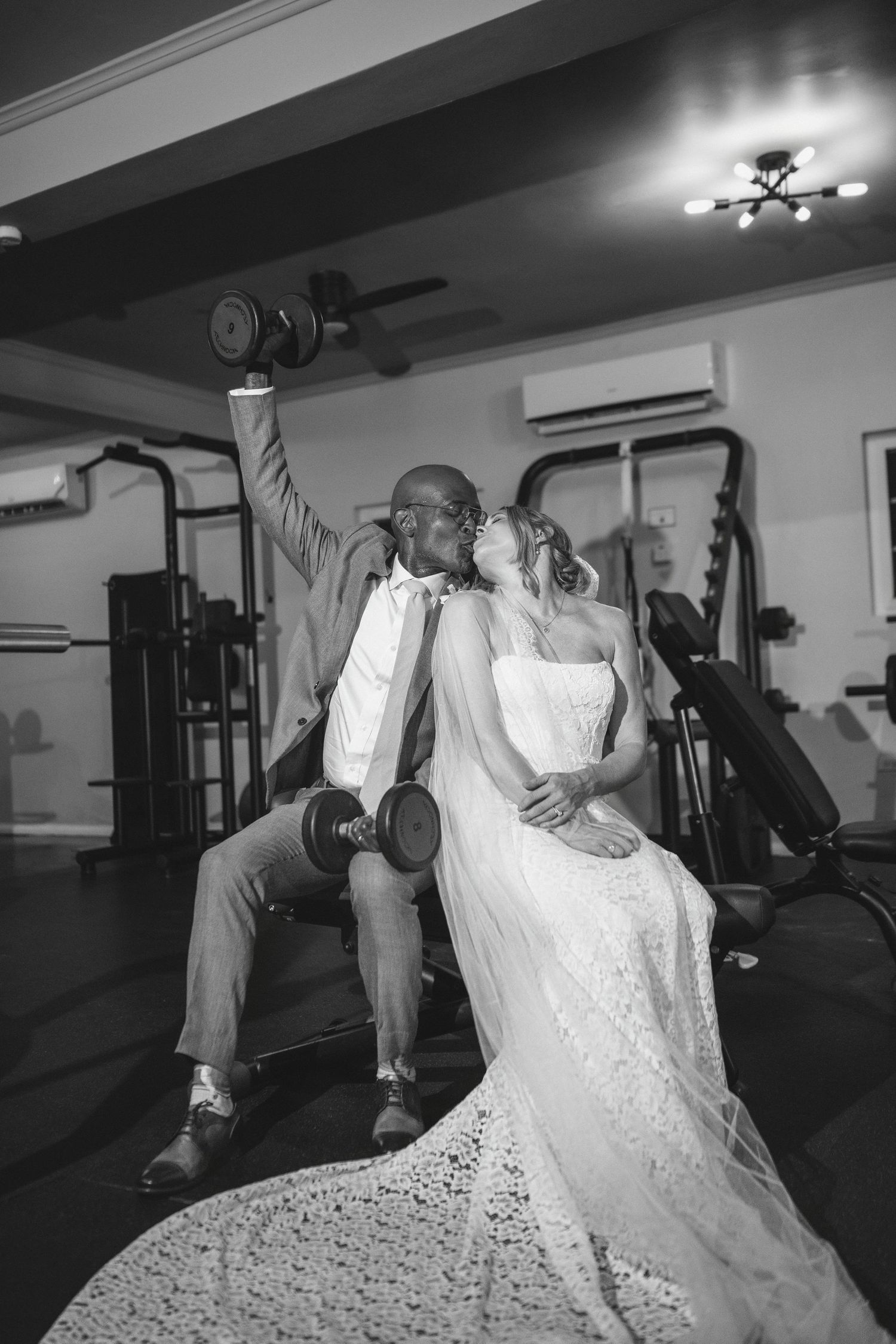 Black and white photo of a couple in wedding attire kissing in a gym, groom lifting dumbbells while bride sits beside him.